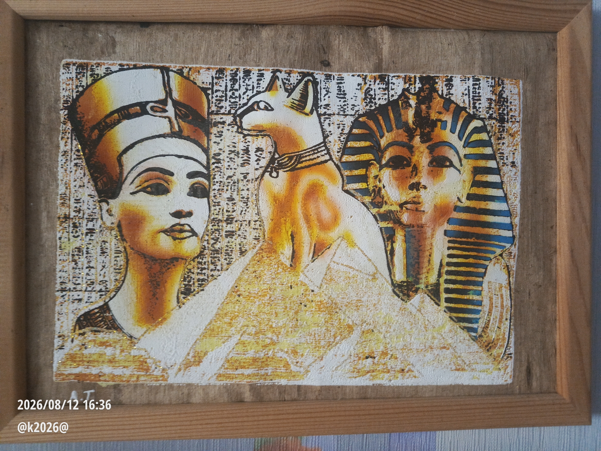The image shows a framed painting featuring ancient Egyptian motifs. The painting depicts the profiles of Nefertiti and a pharaoh, with a stylized cat in the center. The background has a hieroglyphic texture. The painting is on a wooden frame and is hanging on a textured, possibly wooden, wall. A timestamp "2026/08/12 16:36" and a username "@k2026@" are superimposed in the lower left corner. The scene is likely a domestic setting in Minsk, Belarus.