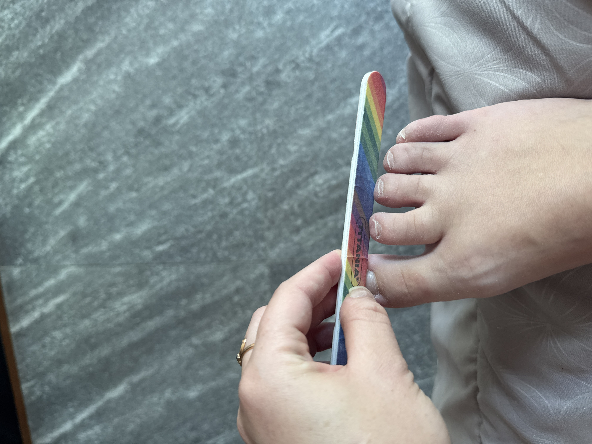 A close-up, high-angle shot captures a person's left foot with the toes filed by a rainbow-striped nail file. The person's hand, adorned with a gold ring, holds the file. The background shows a textured gray surface, likely a floor. The scene is set in Rawai, Thailand.