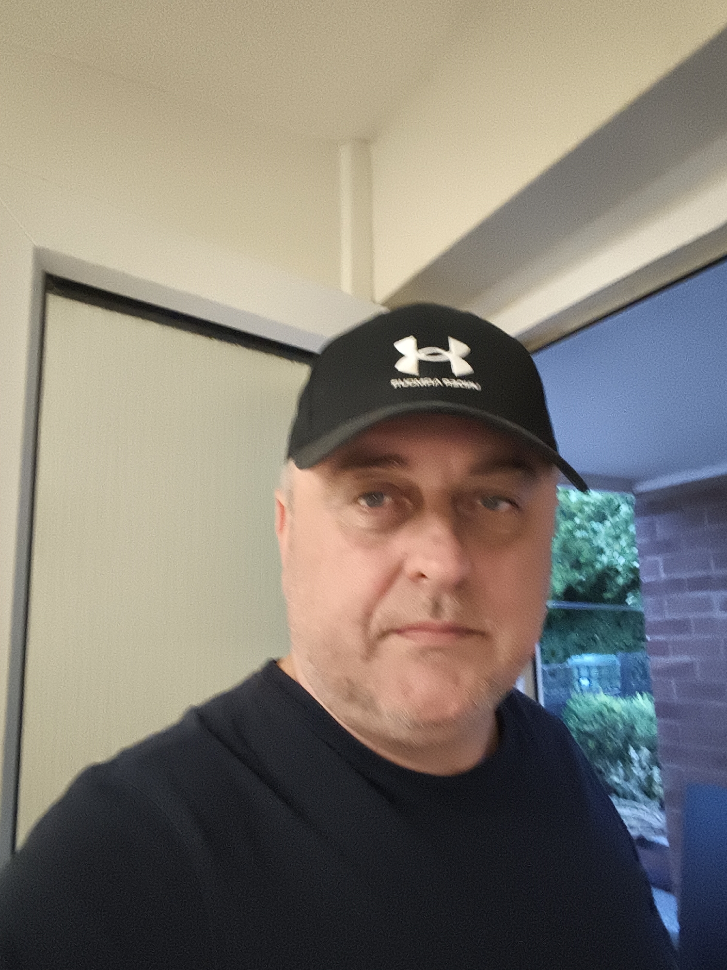 A waist-up portrait captures a middle-aged man looking directly at the camera with a neutral expression. He has light skin, short light hair partially visible, and appears to have light stubble. He is wearing a dark blue or black t-shirt and a black baseball cap.
The cap features a prominent white Under Armour logo (stylized 'UA') on the front, with additional white text below it that appears to read "PHOENIX RISING," although it is slightly obscured and blurry.
The man is positioned indoors, possibly within a doorway or in front of a large window. To his left, a light-colored wall or doorframe is visible. The ceiling above him is also light-colored, featuring a white beam or trim. To his right, an outdoor scene is visible through the opening, showing dark green foliage (likely trees or bushes) and a reddish-brown brick wall, characteristic of residential architecture, consistent with the Huntingdonshire, United Kingdom location. A faint blue hue in the background suggests either the twilight sky or artificial outdoor lighting, indicating the photo was taken at dusk or night.