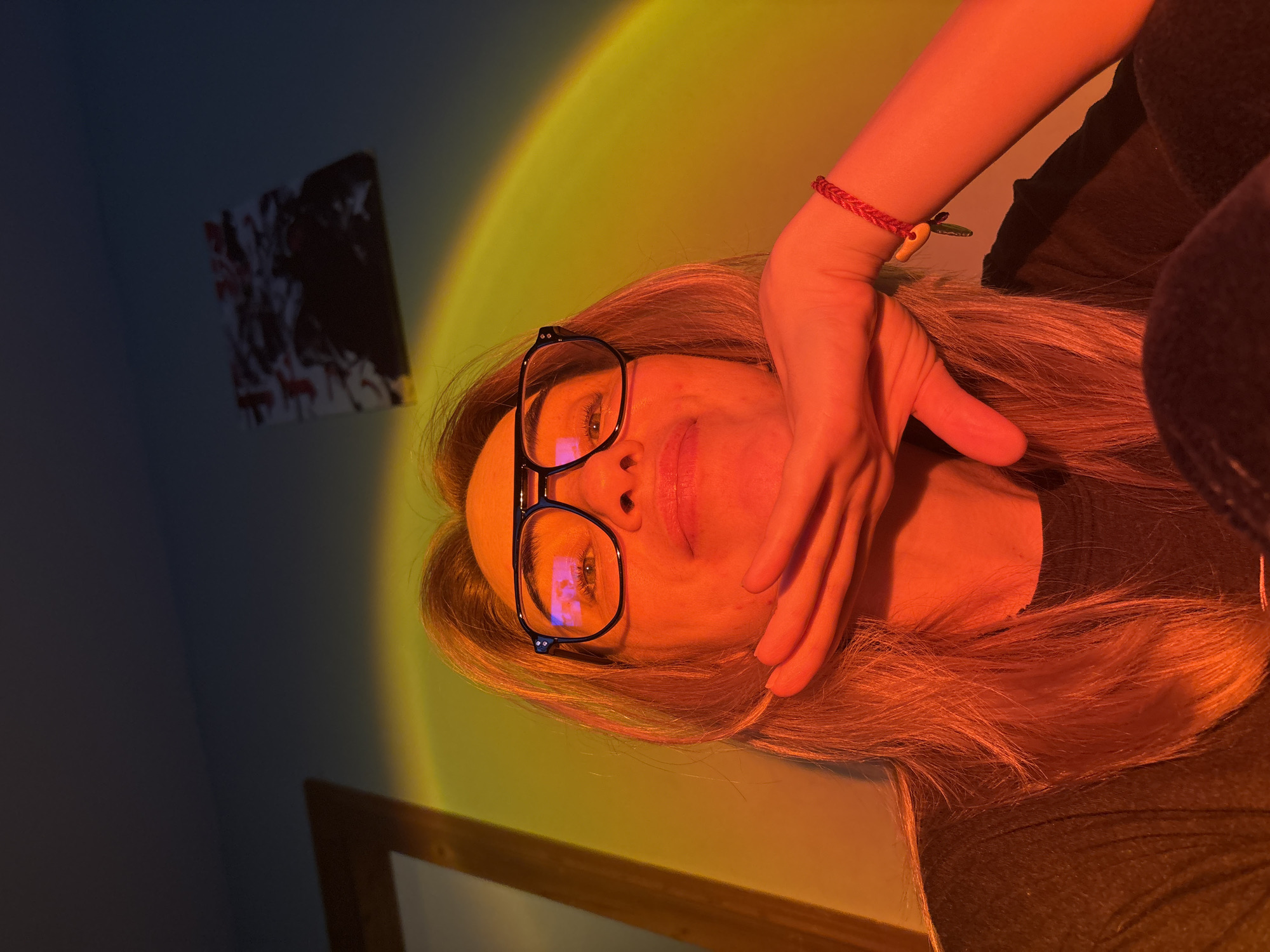 The media file presents an indoor portrait of a woman, oriented sideways. The subject, a woman with long, light-colored hair, is positioned with her head tilted, and her face partially obscured by her hand. She wears dark-framed glasses, through which her visible eye looks calmly towards the viewer. On her wrist, she wears a braided red bracelet featuring a small, light-yellow charm, possibly a miniature carrot or similar shape. She is dressed in a dark-colored top.
The background is dominated by an arc-shaped, gradient light projected onto a wall, creating a vibrant wash of warm yellow and orange on the right, transitioning to cooler blue and purple tones on the left. This distinctive lighting suggests the use of a "sunset lamp" or similar device, casting a strong reddish-orange glow on the woman's hair and the exposed part of her face. To the far left, a dark-toned, framed picture with abstract or chaotic patterns is visible on the dimly lit wall. The bottom of the image shows the edge of a wooden frame or furniture piece. The overall setting is intimate, suggesting a room at night or with controlled artificial lighting.
The activity depicted is a posed photograph. No visible text is present in the image. The location is unknown, Russia.