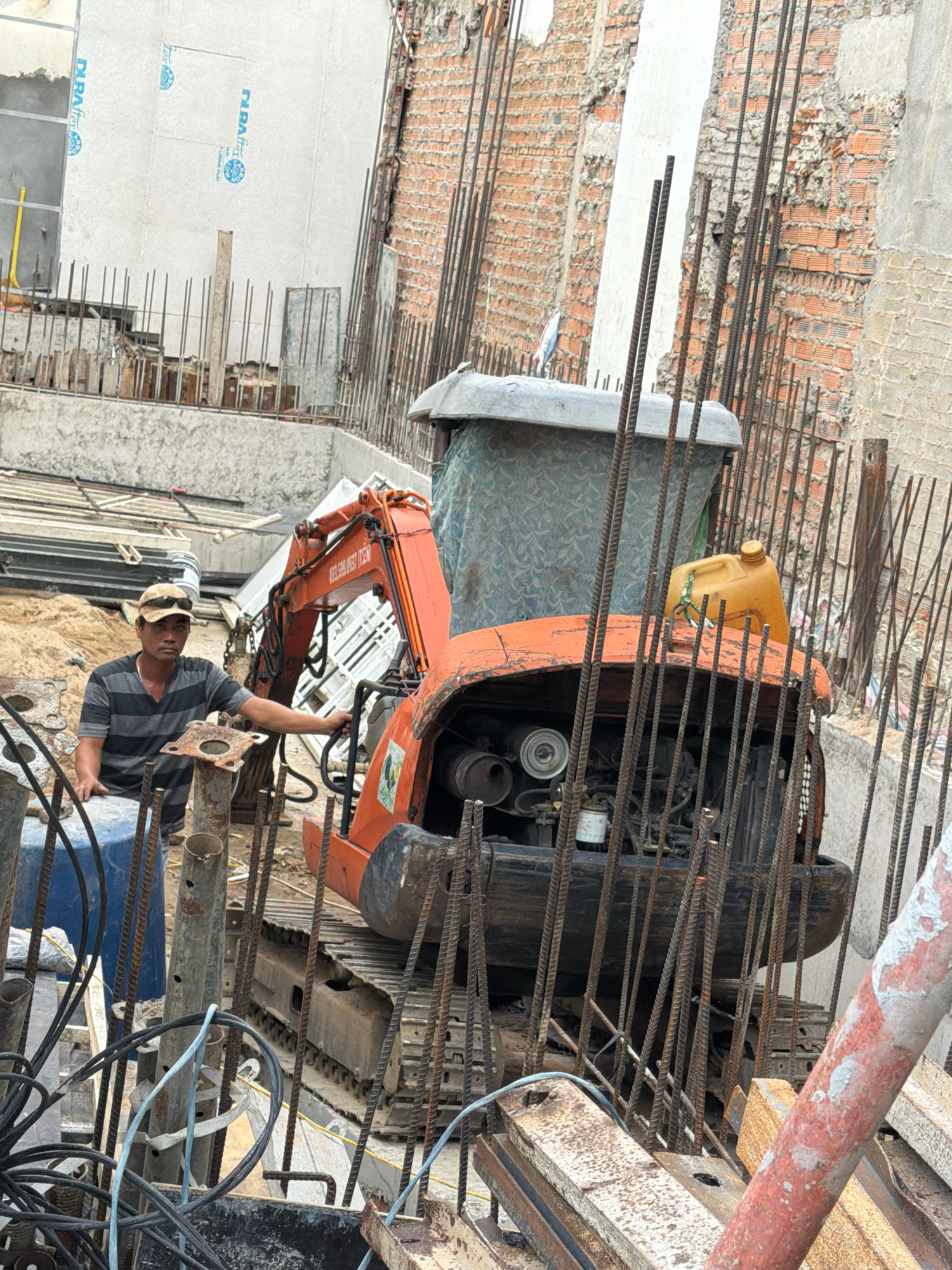 The image shows a construction site in Nha Trang, Vietnam. A man wearing a striped shirt and a cap stands next to an orange mini-excavator. The excavator is surrounded by rebar. There is a yellow plastic container on top of the excavator's engine compartment. The background shows unfinished brick walls and concrete structures. The ground is covered in sand and debris.