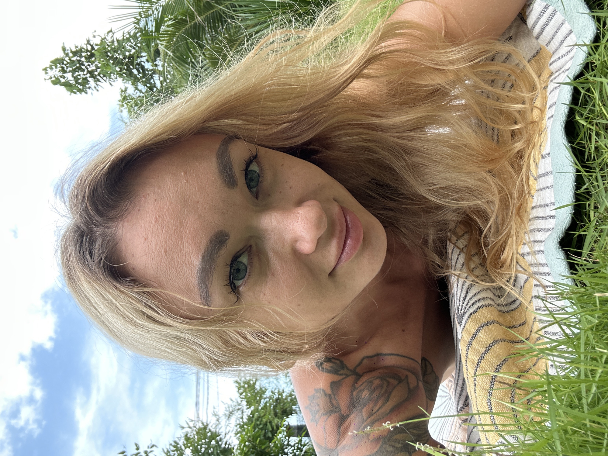 An outdoor, upward-angled portrait captures a woman with long, wavy blonde hair lying on her side on a striped towel placed on green grass. Her head is positioned towards the left, and she looks directly at the camera with a subtle, calm smile. She has light skin, green eyes framed by visible eyelashes, and a prominent floral tattoo depicting a rose and leaves on her right shoulder and upper arm.
Her blonde hair spreads out across the towel, which features stripes in yellow, white, and light blue with thinner black lines. The background reveals a clear blue sky with white clouds and abundant green foliage, including discernible palm fronds and other tropical plants. The bright lighting suggests it is daytime, likely sunny. The image has a relaxed, natural feel, consistent with an outdoor setting in a warm climate like Mũi Né, Vietnam. No text is visible in the media.