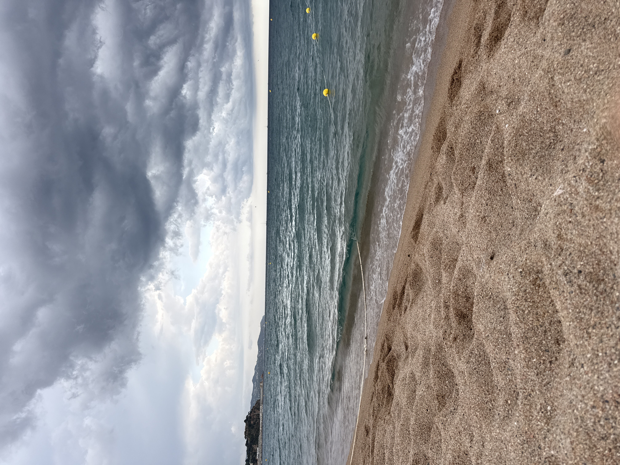 A beach scene in Blanes, Spain, under a dramatic, cloudy sky. The foreground is dominated by wet sand with visible footprints, leading towards the ocean. Gentle waves are washing ashore. In the mid-ground, the dark blue sea stretches to the horizon, marked by a series of yellow buoys on a rope, likely delineating a swimming area. In the distance, a hilly coastline with buildings and vegetation can be seen. The overall atmosphere is moody due to the heavy cloud cover.