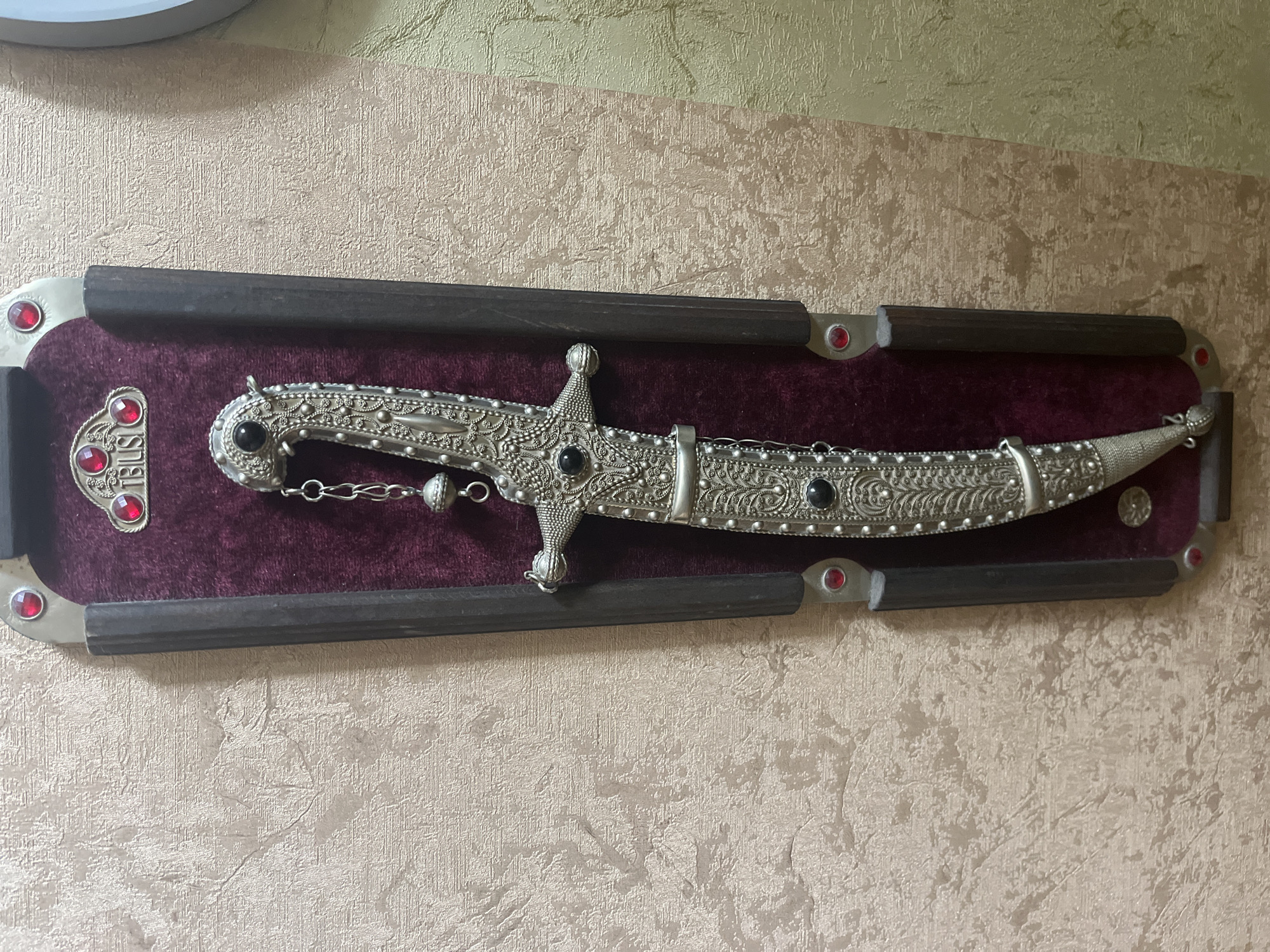 The image displays a decorative dagger mounted on a wall in Vladimirovka, Russia. The dagger is ornate, with intricate silver detailing and black stones, and is presented within a dark wooden frame lined with burgundy velvet. The frame is adorned with red jewel-like accents at the top and bottom corners. The wall behind the display has a textured, light-colored wallpaper. The lighting is even, and the focus is sharp on the dagger and its display. There are no people or animals visible in the scene. The image is a static, detailed close-up of the decorative item.