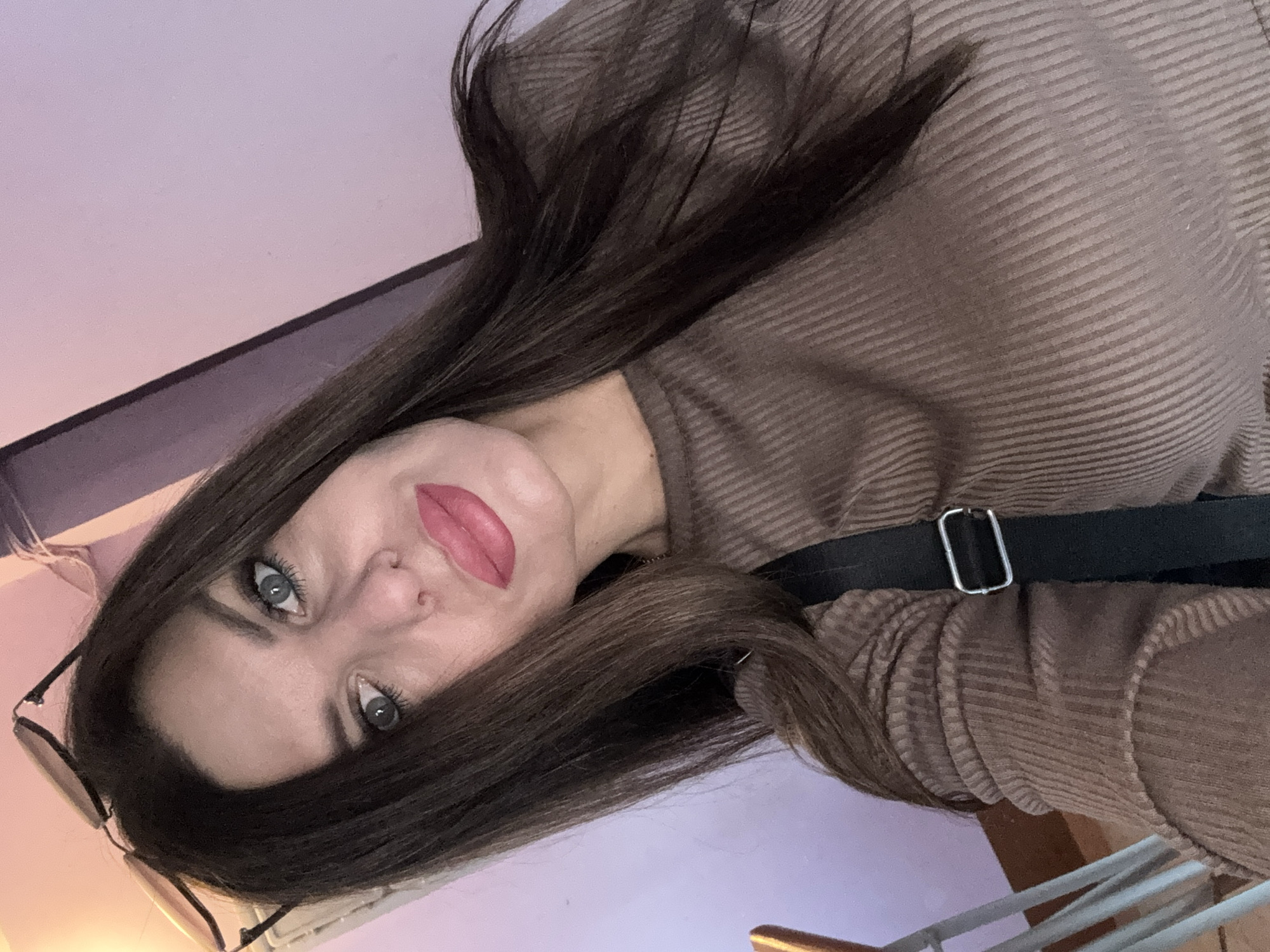 A woman with long, dark brown hair and light green eyes is taking a selfie. She is wearing a brown, ribbed, long-sleeved shirt and a black strap across her chest. Her lips are painted with a pink lipstick. Sunglasses are perched on top of her head. The background appears to be an indoor setting with a light purple wall and a wooden railing. The scene is consistent with a casual selfie taken in a residential or public indoor space in Chaikovsky, Russia.