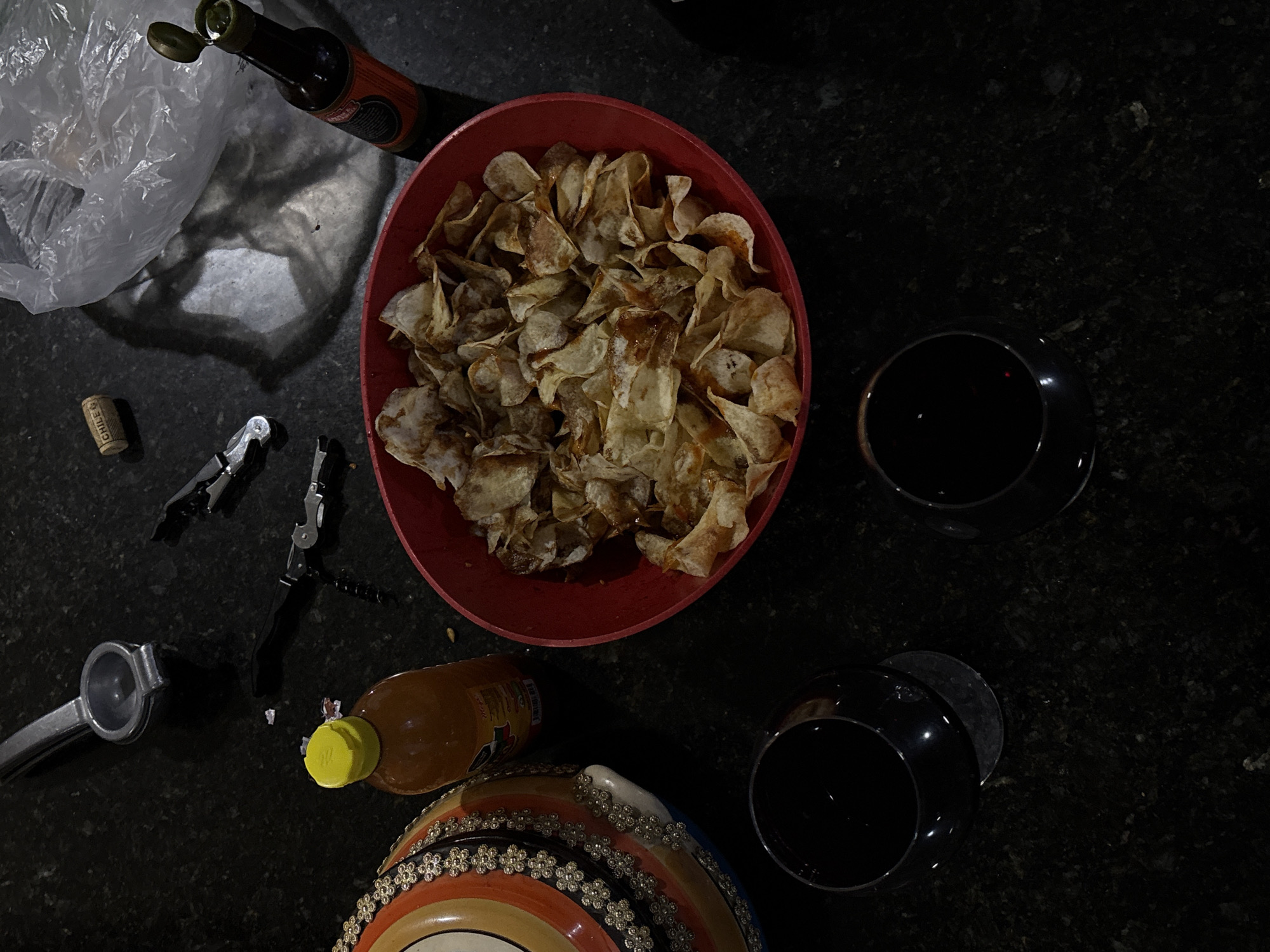 The image depicts a dark countertop setting. A red bowl is filled with potato chips, some of which appear to be drizzled with a dark sauce. Beside the bowl are two wine glasses filled with a dark liquid, presumably red wine. A wine opener, a cork, a plastic bag, a bottle of hot sauce, and a citrus juicer are scattered around. A decorative item is partially visible at the bottom of the frame. The location is Santa Clara Ocoyucan, Mexico, but there is no information to confirm this. There is no evidence of people or any specific actions or interactions. The environment seems to be indoors, likely in a kitchen or dining area.