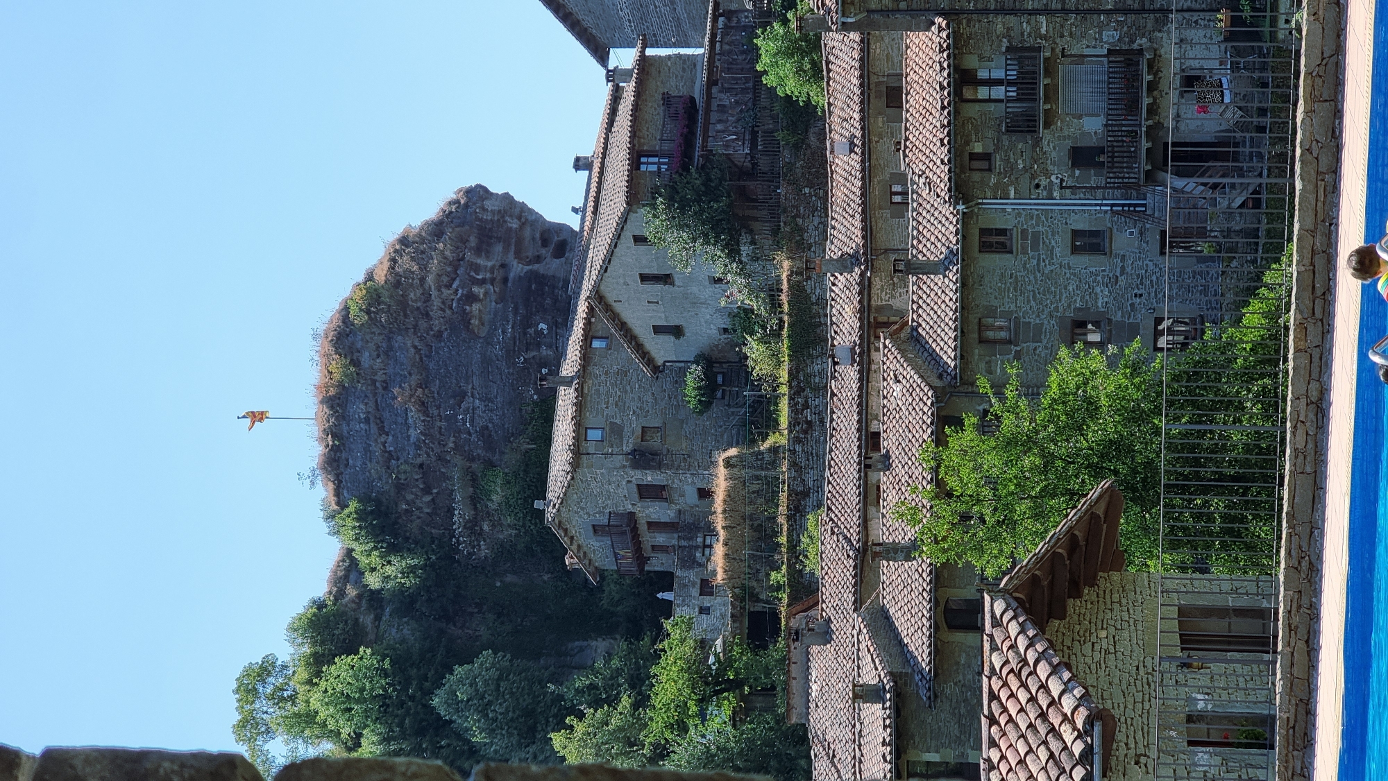 The image shows a view of the town of Rupit i Pruit, Spain, featuring traditional stone houses with red tile roofs nestled into a hillside. A large, weathered rock formation dominates the upper background, topped with a flag. Lush green trees and vegetation surround the buildings and the rock. In the foreground, a bright blue swimming pool occupies the bottom portion of the frame, with a glimpse of two people, one child and one adult, sitting on the edge of the pool. The scene depicts a sunny day with a clear blue sky.