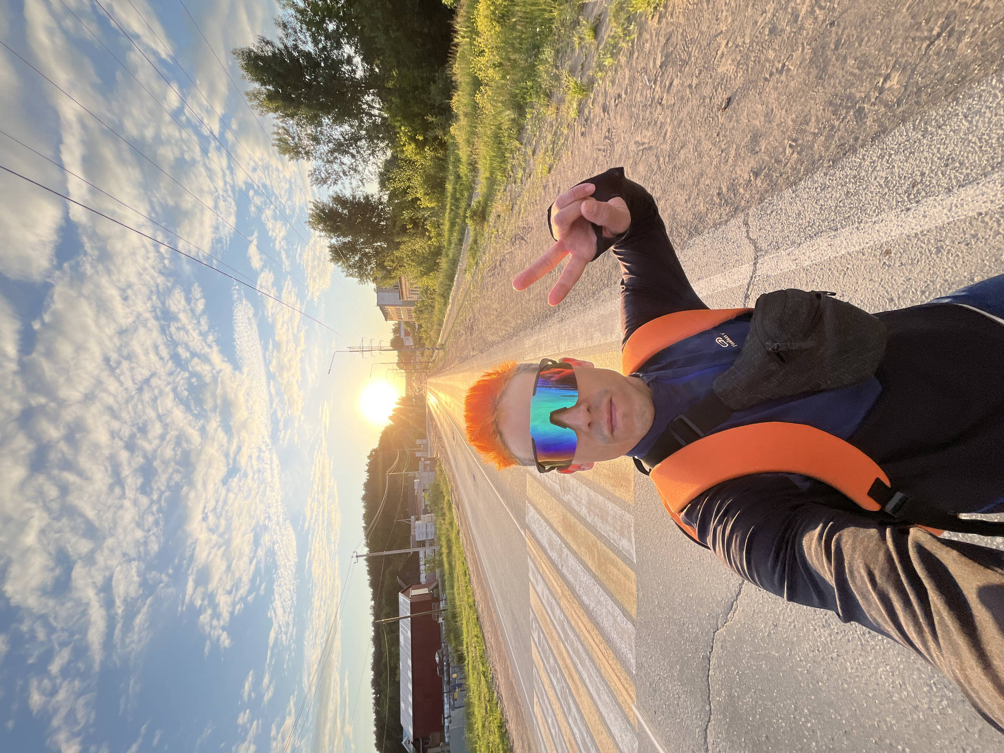 An adult male with bright orange hair styled upwards and wearing large, multi-colored reflective sports sunglasses is positioned in the foreground, taking a selfie. He wears a dark long-sleeved top, an orange backpack strap over his right shoulder, and a black fanny pack across his chest. His right hand is raised, forming a peace sign (V-sign) with index and middle fingers extended, palm facing away, and covered by a black sleeve or glove. A partial "S. Italian" logo is visible on the backpack strap.
The man stands on or near an asphalt road featuring a pedestrian crosswalk with white and faded yellow stripes in the foreground. The road extends straight into the distance, aligning directly with the low-lying setting sun, which casts a strong golden light across the scene.
To the left of the road, a grassy verge leads to a red-roofed building resembling a barn, with other structures and construction materials visible nearby. To the right, a dirt shoulder and a strip of grass are bordered by numerous mature trees, with additional buildings partially visible further down. Overhead, multiple power lines stretch across the sky, supported by utility poles. The sky is largely clear blue with scattered white, wispy clouds. The scene is illuminated by the intense golden glow of the sunset, creating distinct highlights and long shadows. The location is an outdoor road environment in Chaikovsky, Russia.