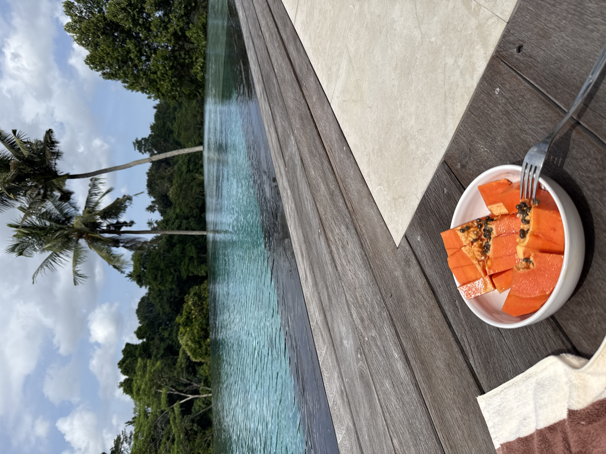 The image depicts a scene in Ubud District, Indonesia, showcasing a bowl of papaya on a wooden surface near an infinity pool. The papaya, sliced and containing seeds, sits in a white bowl with a fork placed inside. A towel rests alongside the bowl. The wooden surface appears to be part of the pool deck, which adjoins a tiled area. The infinity pool extends towards a lush green landscape with palm trees, under a partially cloudy sky. The environment suggests a tropical resort or private villa setting.