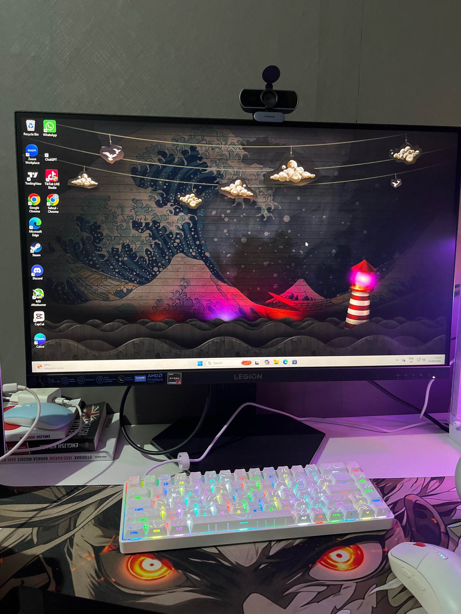 This is a close-up, eye-level, indoor, still photograph of a gaming setup. The primary subject is a desktop computer monitor displaying a stylized, dark, Japanese-inspired wallpaper featuring a large wave, mountains, and a lighthouse. The monitor is positioned on a white desk. To the left of the monitor are several icons for various applications, including Recycle Bin, WhatsApp, Zoom, ChatGPT, TradingView, TikTok LIVE Studio, Google Chrome, Microsoft Edge, Steam, Discord, MSI Afterburner, CapCut, and Canva. A webcam is mounted on top of the monitor.
Below the monitor, on the desk, are various computer peripherals. A white mechanical keyboard with RGB backlighting is prominently displayed, its keys illuminated with a rainbow of colors. A white computer mouse is visible to the right of the keyboard. To the left of the keyboard, a stack of books is visible. The monitor stand has "LEGION" branding. The desk surface is covered by a large gaming mousepad featuring an anime-inspired illustration of a fierce, glowing eye. The background appears to be a wall with a textured finish. The lighting in the scene is a mix of ambient light and the purple glow from the monitor and possibly other RGB lighting elements, creating a vibrant and colorful atmosphere. There are no people present in the image. The overall scene suggests a personal computer workstation focused on gaming or content creation.
The image does not provide any specific visual cues to indicate its location in Cimahi, Indonesia.