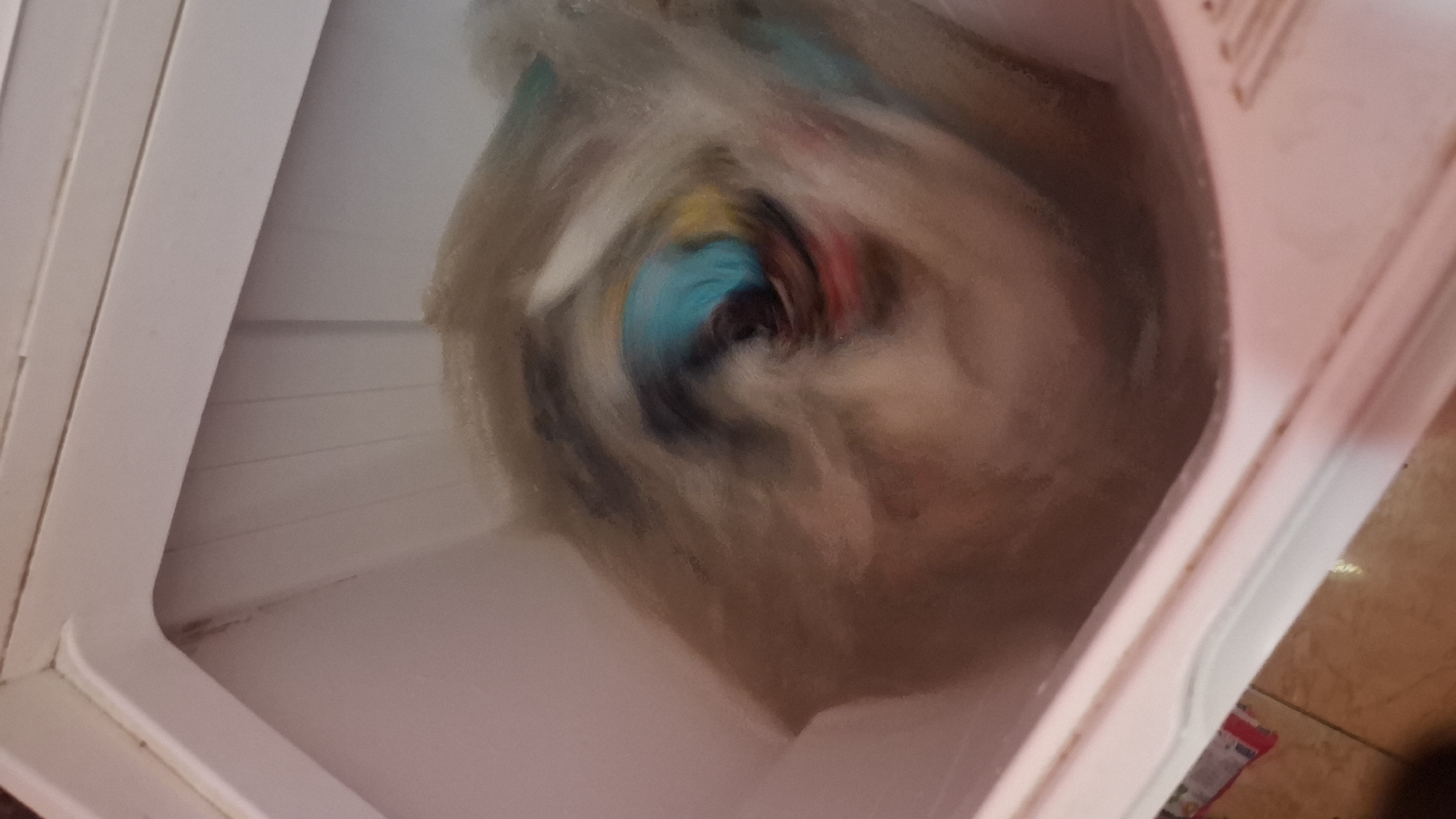 The image shows the interior of a white washing machine, viewed from above. The drum of the washing machine is filled with clothes that are blurred due to motion. The clothes appear to be a mix of colors, with some prominent shades of blue and red. The washing machine is located in a residential setting, with the floor and part of a cabinet visible in the lower left corner of the image. Based on the context provided, this scene is located in the city of Cibinong Hilir, Indonesia.