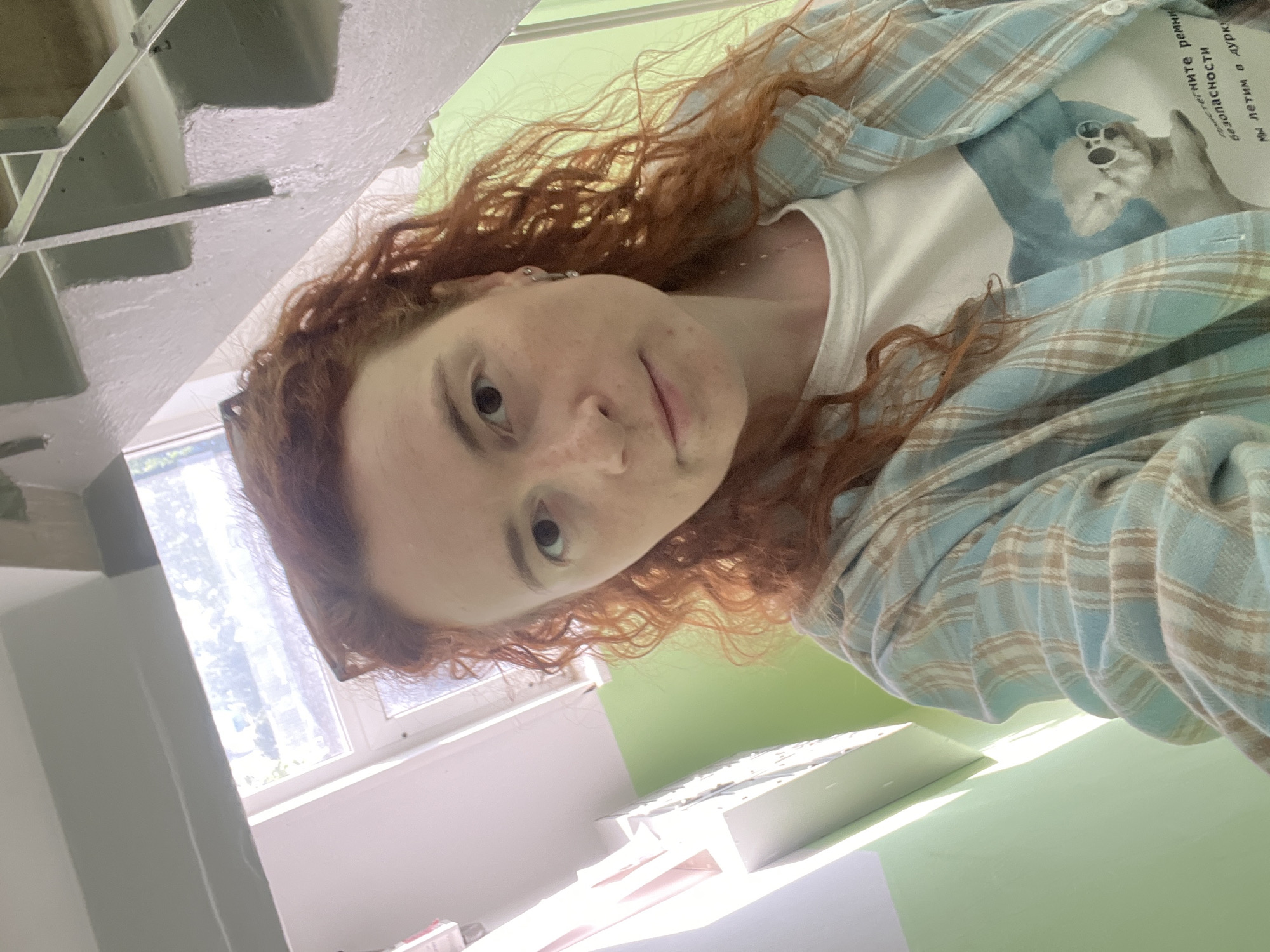 A pale-skinned, red-haired woman is pictured in an indoor environment. She is wearing a white t-shirt with a graphic image, partially covered by a blue and tan plaid shirt. The setting appears to be a stairwell, as indicated by the steps visible on the left side of the image. A window with natural light is positioned to the left of the woman. The background shows what seems to be a light green wall and a desk, suggesting that the location may be within a building in Chaikovsky, Russia.