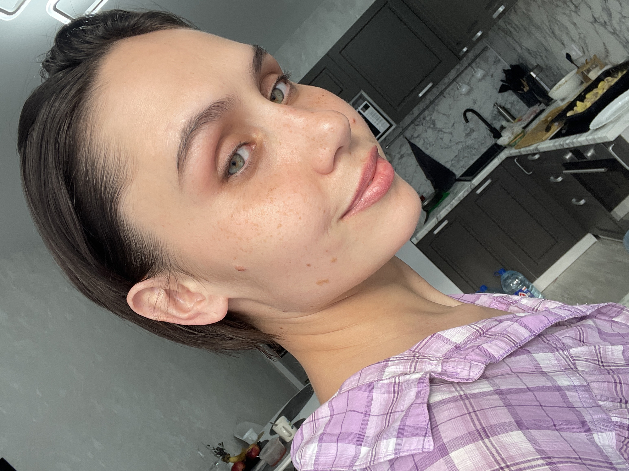 A medium shot captures a fair-skinned individual with short, dark brown hair pulled back from their face, positioned indoors against a kitchen backdrop. The person has freckles across their nose and cheeks, light green eyes, and subtly glossy lips. They are wearing a purple and white plaid shirt. The kitchen features dark cabinets, a marble-patterned backsplash, and a sink area with a black faucet and various kitchen utensils and dishes. A water bottle is visible on the floor. The location in the city of Izhevsk, Russia cannot be determined from the image.