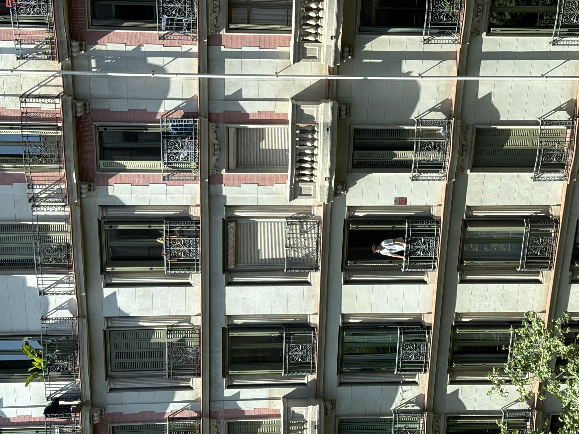 The image depicts the exterior of a multi-story building in Barcelona, Spain, characterized by ornate window railings and a symmetrical facade. A person, wearing a white shirt, is standing in one of the windows. The building features a light-colored exterior with pink brick accents. Some windows have shutters that are either closed or partially open. A tree is visible on the right side of the building. A straight, linear object runs across the facade.