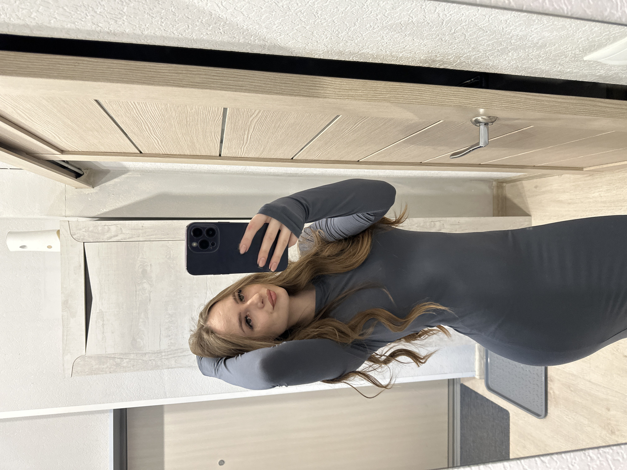 A woman is taking a selfie in what appears to be a bathroom. She is wearing a long-sleeved grey dress. Her hair is long, blonde, and wavy. She is holding a black smartphone in her right hand to take the photo. The background shows a bathroom mirror, a light-colored vanity cabinet, a toilet paper dispenser, and a glimpse of the floor. The location is Unknown, Unknown.