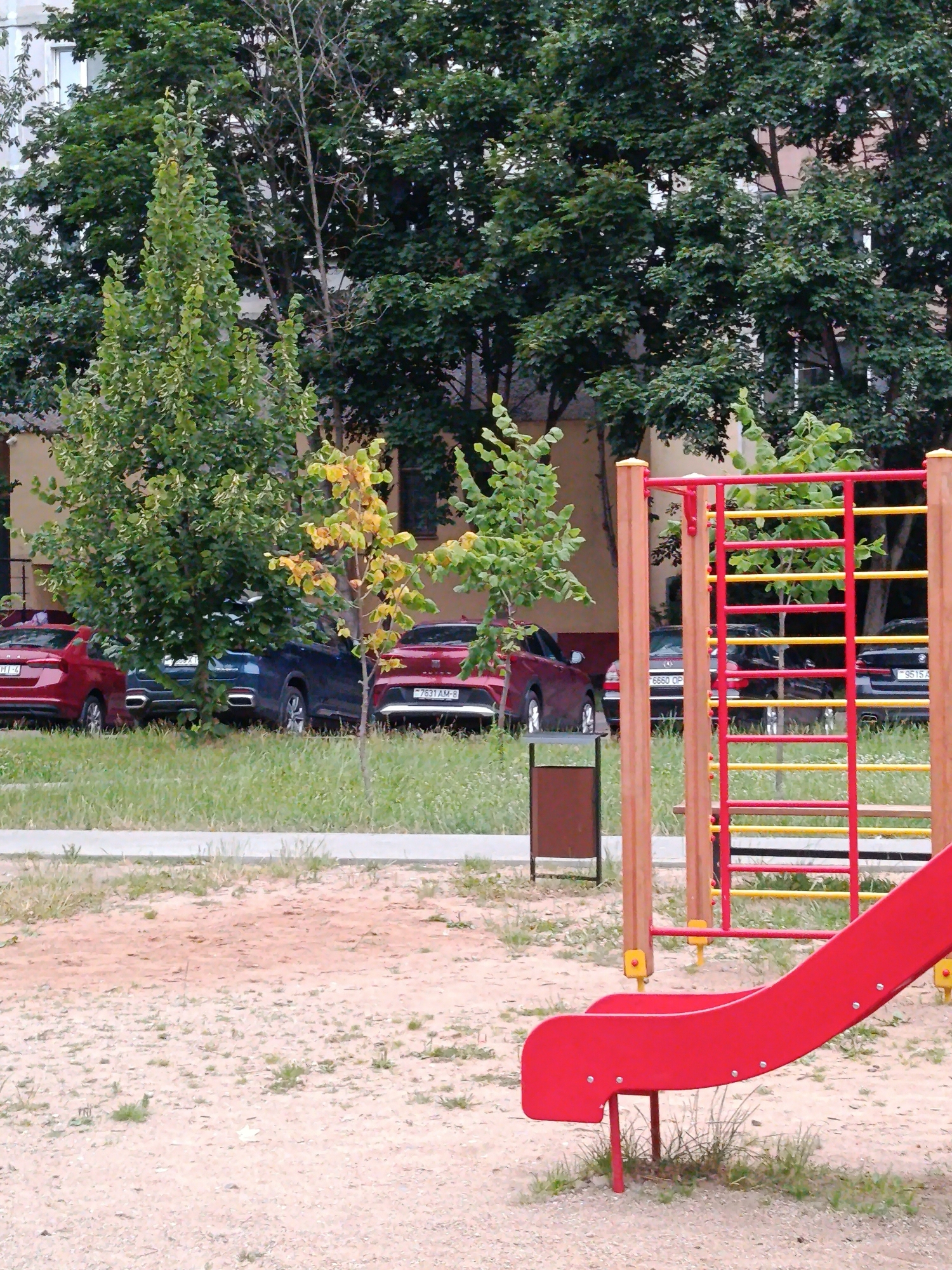 This is a medium-shot, vertically oriented, outdoor photograph taken during the daytime in what appears to be a residential area. The foreground features a section of a red plastic slide from a playground, with sandy ground and patches of sparse grass beneath it.
Behind the playground equipment, a line of parked cars is visible, partially obscured by trees and grass. The cars are of various colors, including red, dark blue, and black. License plates are visible on some of the cars, with text such as "7531 AM-8", "6660 OP", and "9515 A".
A dense canopy of green trees dominates the middle ground and background, with some trees showing hints of yellowing leaves, suggesting the transition into autumn. Behind the trees, a light-colored apartment building is visible, with several windows. The overall scene depicts a typical urban residential setting with a playground and parked vehicles, set against a backdrop of mature trees. The lighting suggests it is a bright day, but there are no strong shadows. The weather appears to be mild. There are no people visible in the photograph.