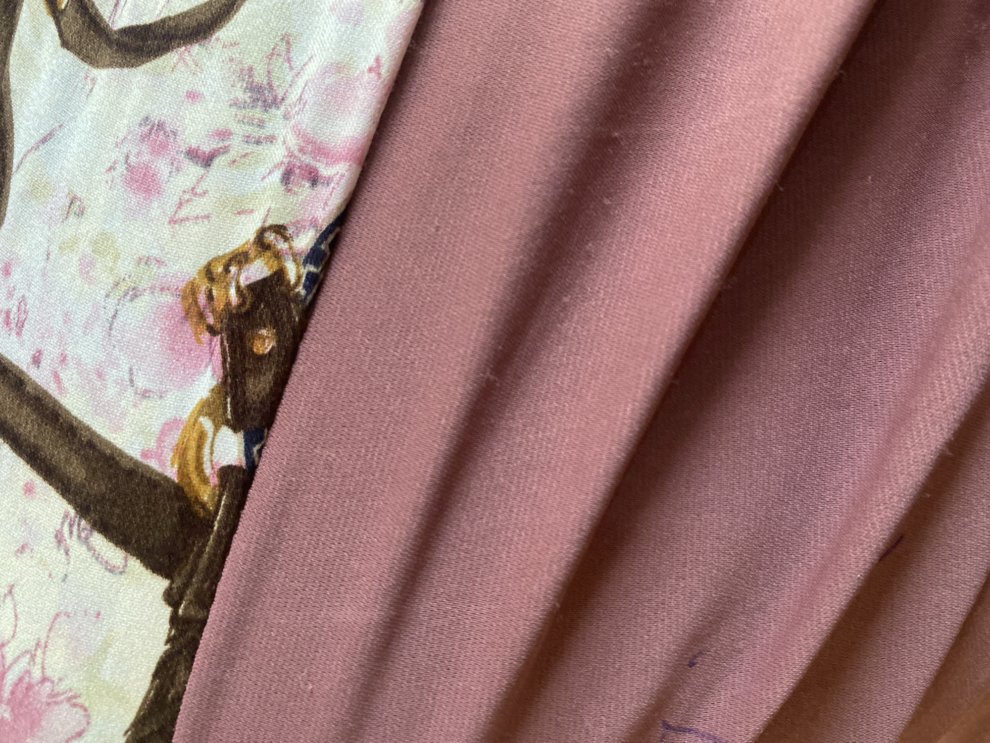 The image displays a close-up view of two pieces of fabric. The top fabric features a pattern of pink cherry blossoms and dark brown branches. It is laid on top of a plain, dusty rose-colored fabric, which appears to be creased and folded. The lighting suggests an indoor setting. There is no indication of the city of Jauro Yinu, Nigeria, within the image.