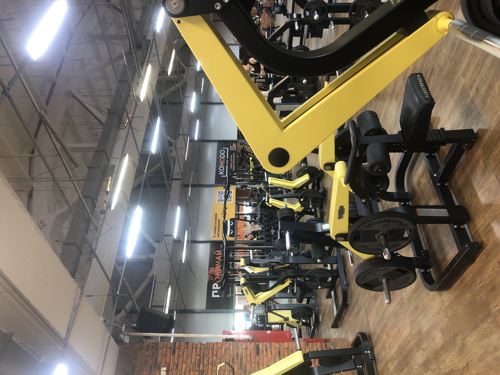 Here's a description of the image:
The image shows the interior of a gym, likely located in Chaikovsky, Russia, based on the Cyrillic text on a banner. The gym is filled with exercise equipment, predominantly black and yellow weight machines. Many of the machines are TECHNOGYM branded. The gym floor is a light wood. The building has a high ceiling with exposed structural beams and fluorescent lighting fixtures. One wall is red brick.