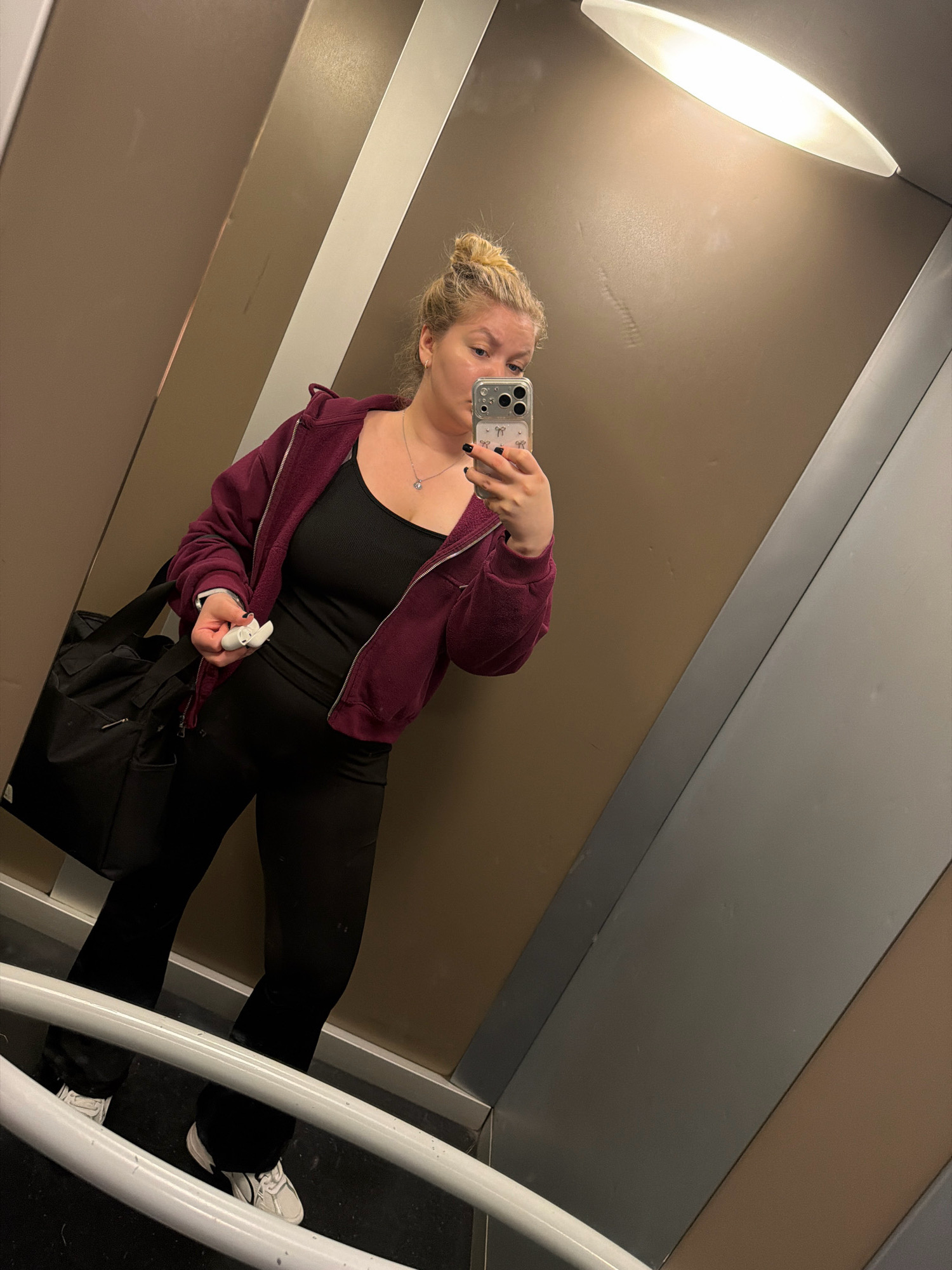 A woman is taking a mirror selfie in what appears to be an elevator. She has blonde hair styled in a bun and is wearing a maroon zip-up hoodie over a black tank top and black pants. She is also wearing white sneakers. She is holding a white object, possibly an earbud case, in her left hand and her smartphone in her right hand. A black duffel bag is slung over her left shoulder. The elevator walls are a mix of brown and metallic grey panels, with a fluorescent light fixture visible on the ceiling. The floor of the elevator is dark and textured, with a white handrail running horizontally across the lower part of the image. The woman's reflection is visible in the mirror. The city of Koszalin, Poland is not visible in the image and cannot be inferred.