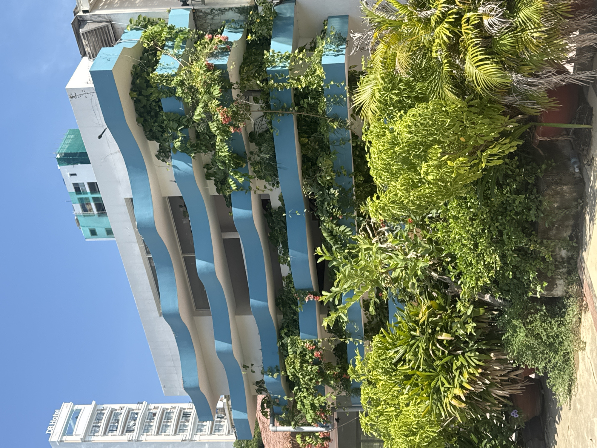 In Nha Trang, Vietnam, a modern building with multiple balconies overflowing with lush greenery stands under a clear blue sky. The building features a distinct wavy, blue trim on each balcony, creating a unique architectural style. In the background, a tall, slender white building with the inscription "DB Hotel" is visible. Construction netting covers the upper floors of another building to the right of the DB Hotel. In the foreground, dense tropical plants and trees in large pots partially obscure the lower levels of the building. The scene is bathed in bright sunlight, suggesting a warm day. There are no people visible in the image.