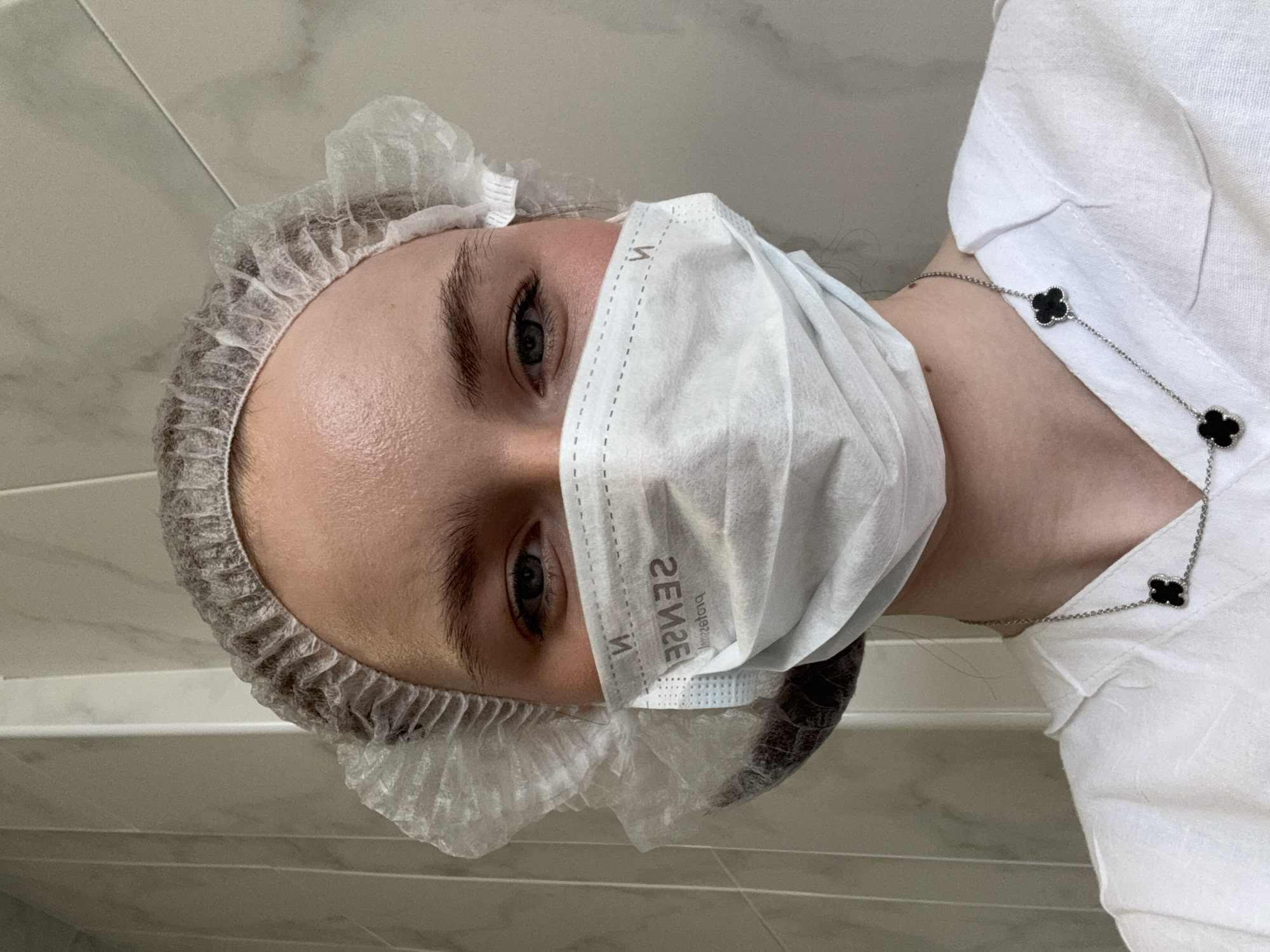 A woman wearing a disposable surgical cap and a white surgical mask with "SENSE" printed on it is the main subject. Her eyes are visible above the mask. She is wearing a white shirt with a silver necklace featuring black clover-shaped pendants. The background appears to be a tiled wall. The image is rotated, giving the impression of an angled perspective. There is no evidence to determine the location in Saint Petersburg, Russia.