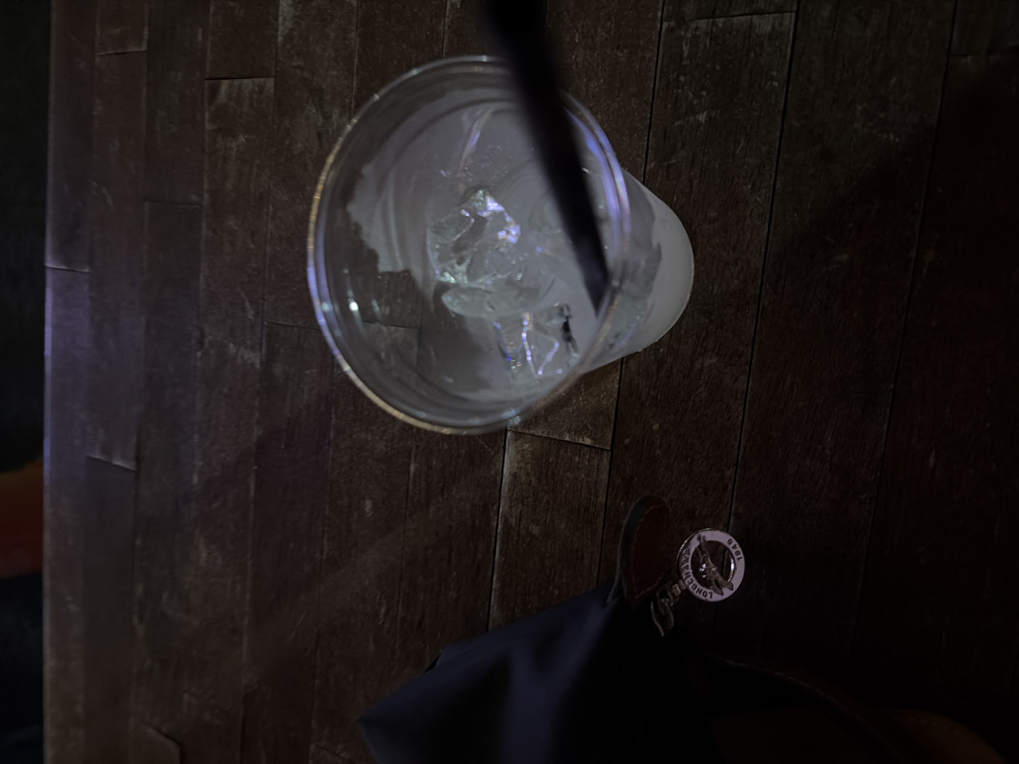 The image depicts a close-up, top-down view of a dark, possibly wooden surface with a disposable plastic cup filled with ice and a straw. Adjacent to the cup, a handbag or tote bag is situated, featuring a Longchamp keychain. The overall lighting is dim and seems to be indoors. There's no readily identifiable information that would specify a location in Austin, United States of America.