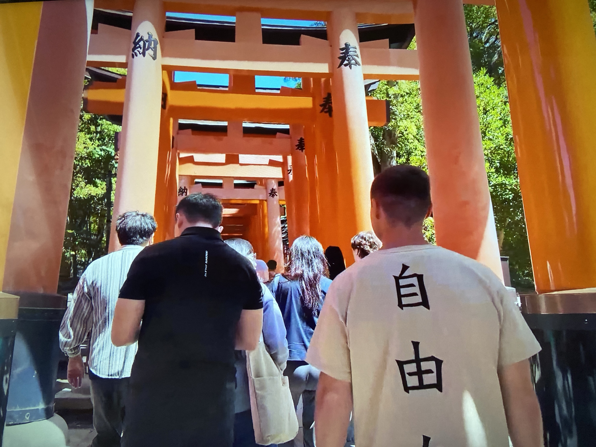 The image shows a group of people walking through the Fushimi Inari Shrine in Kyoto, Japan. The scene is dominated by the numerous orange torii gates that line the path. The people are walking away from the viewer, their backs visible. One person wears a shirt with Japanese characters on the back. The environment consists of trees on either side of the path, partially visible between the torii gates. The overall lighting is bright.