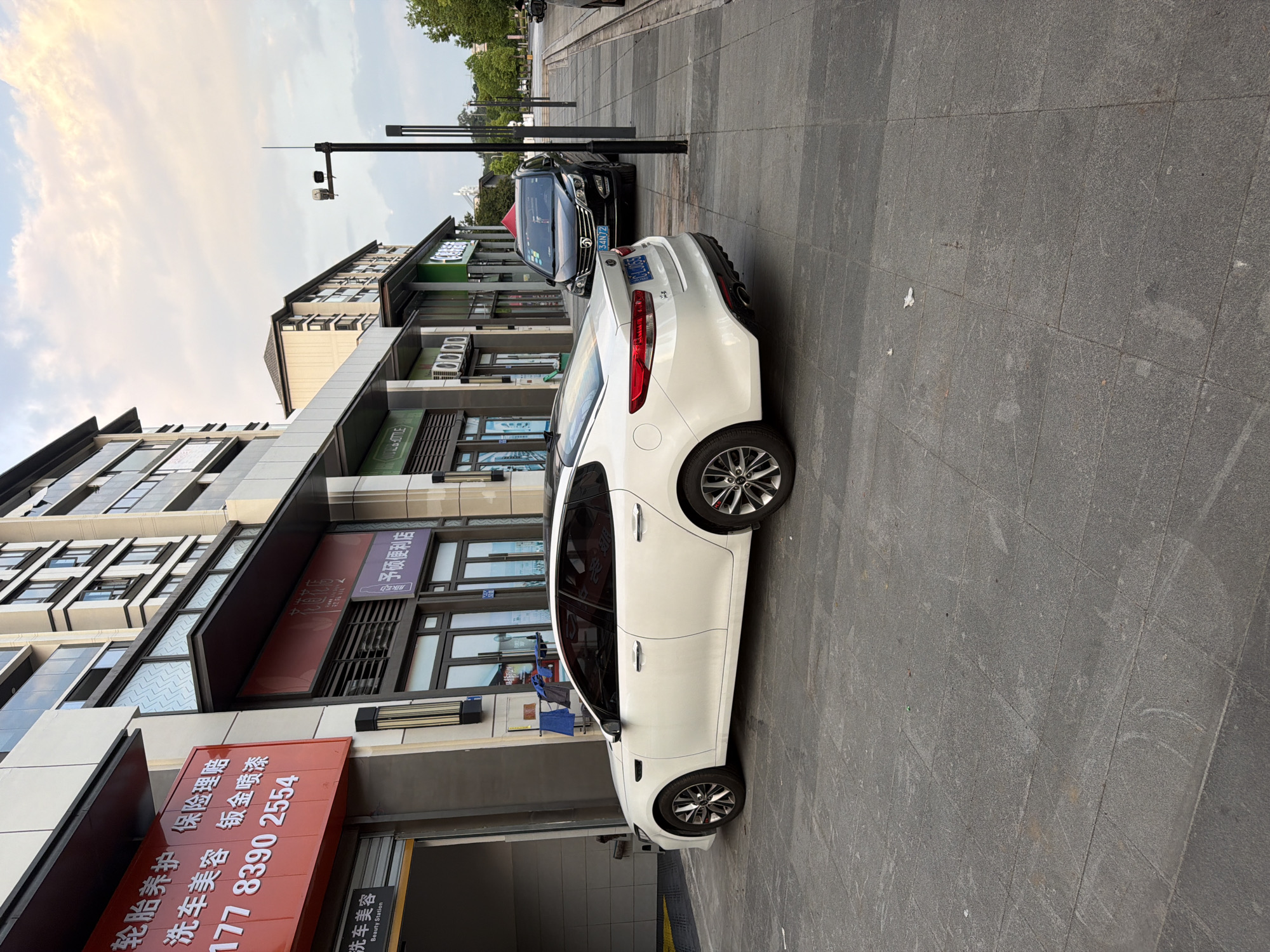 A white Kia Optima sedan and a black SUV are parked on a paved sidewalk in front of a modern building with multiple businesses. The building has large glass windows and awnings over the storefronts. Signs in Chinese are visible on the building. The sky is cloudy with hints of sunset colors. In the background, more buildings and trees are visible. The scene is located in the city of Jiangjin, China.