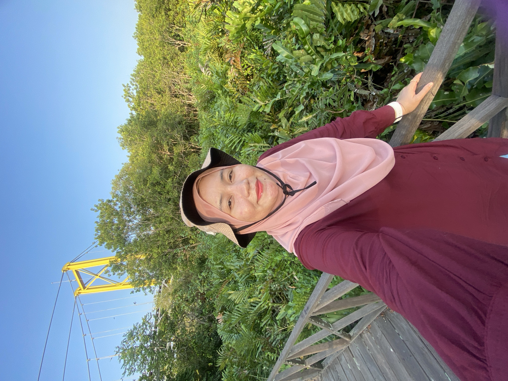 A woman in a pink hijab and a burgundy dress stands on a wooden boardwalk, holding a phone to take a selfie. She wears a wide-brimmed hat. Behind her, lush green foliage stretches out, and a bright yellow suspension bridge tower is visible in the background under a clear blue sky. The scene appears to be in a natural, possibly park-like setting.