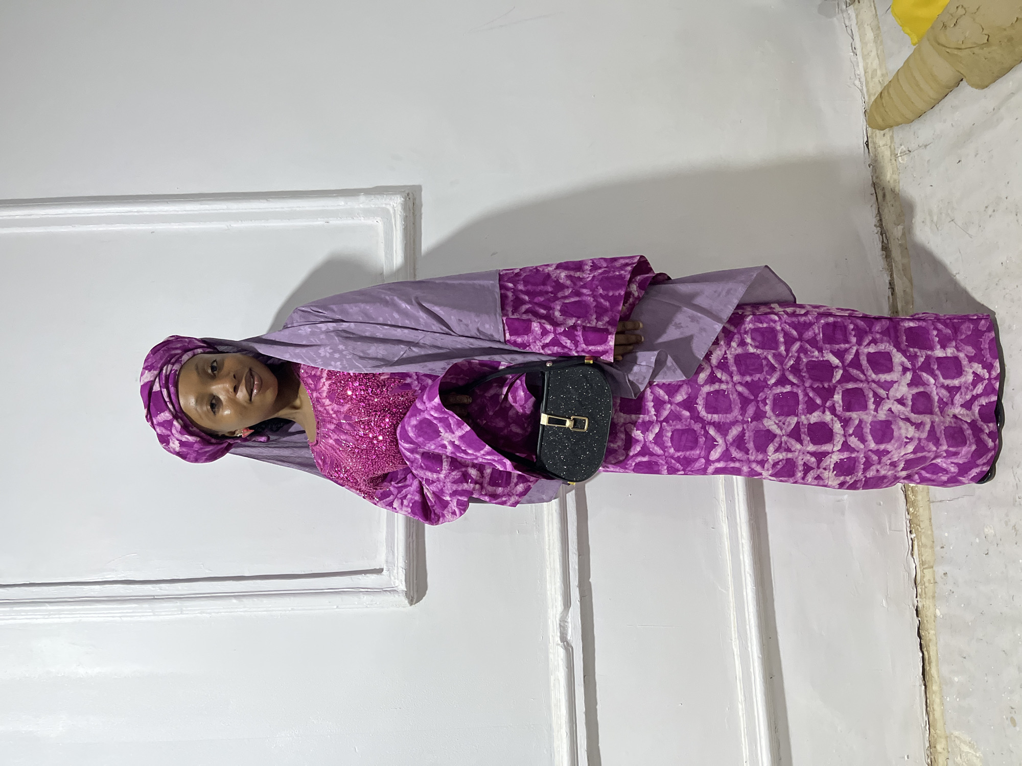 A woman stands against a white wall in Gura Top, Nigeria. She is wearing a purple patterned fabric dress, a matching head wrap, and a pink, sequined top. A black purse hangs from her left shoulder. The wall has a white frame, and the corner of the wall is weathered and discolored. A yellow cloth is visible in the upper right corner of the image.