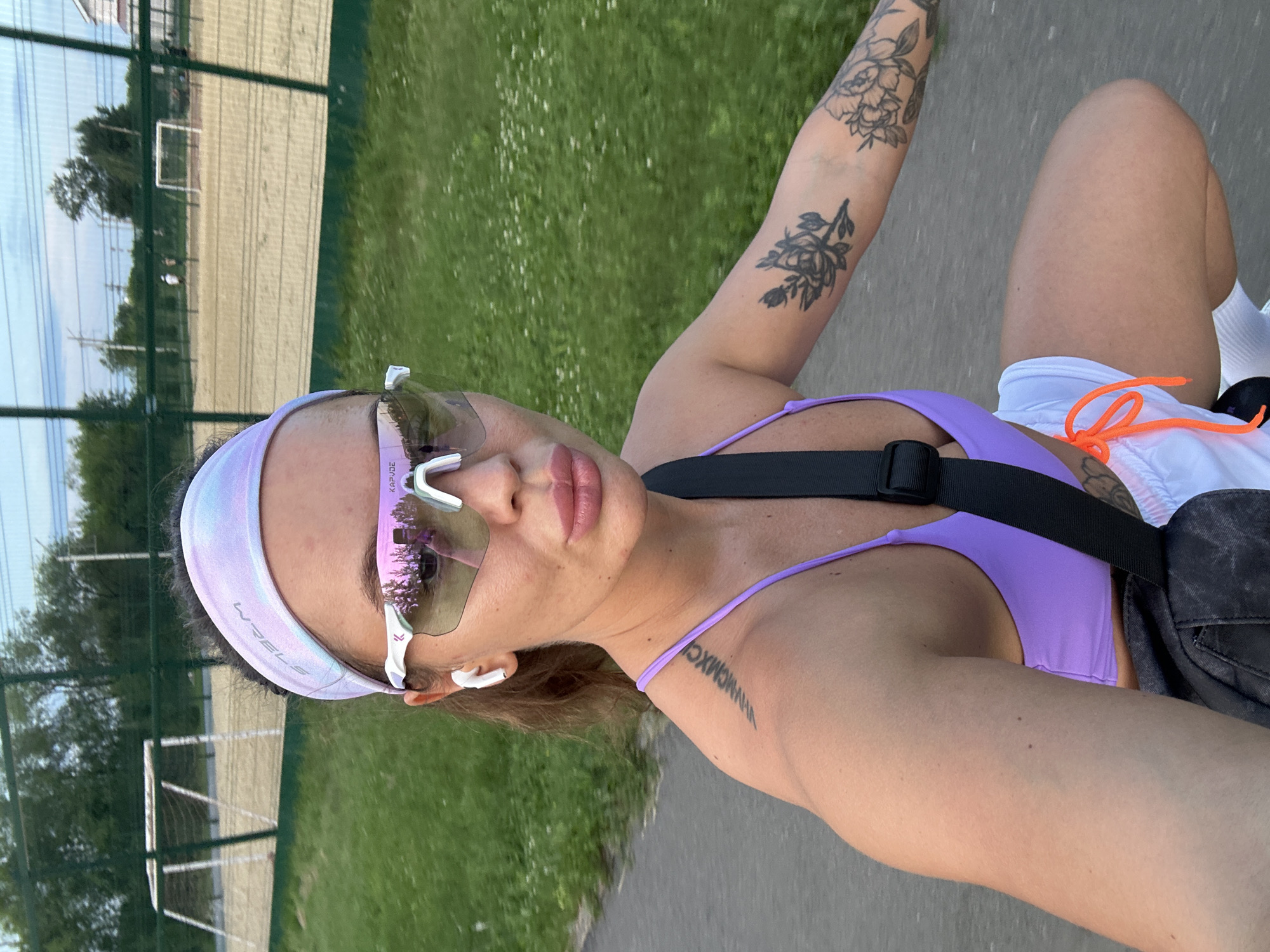 A young woman is pictured from the chest up, riding a bicycle in Yuzhnouralsk, Russia. She is wearing a purple sports bra, white athletic shorts with orange drawstrings, white socks, and athletic shoes. Her hair is pulled back with a tie-dye headband, and she is wearing white wireless earbuds and white athletic sunglasses. She has tattoos on her left arm and shoulder, and a tattoo on her right arm. In the background, there is a soccer field with a goal net, a fence, and trees. The weather is clear, and the sun is shining.