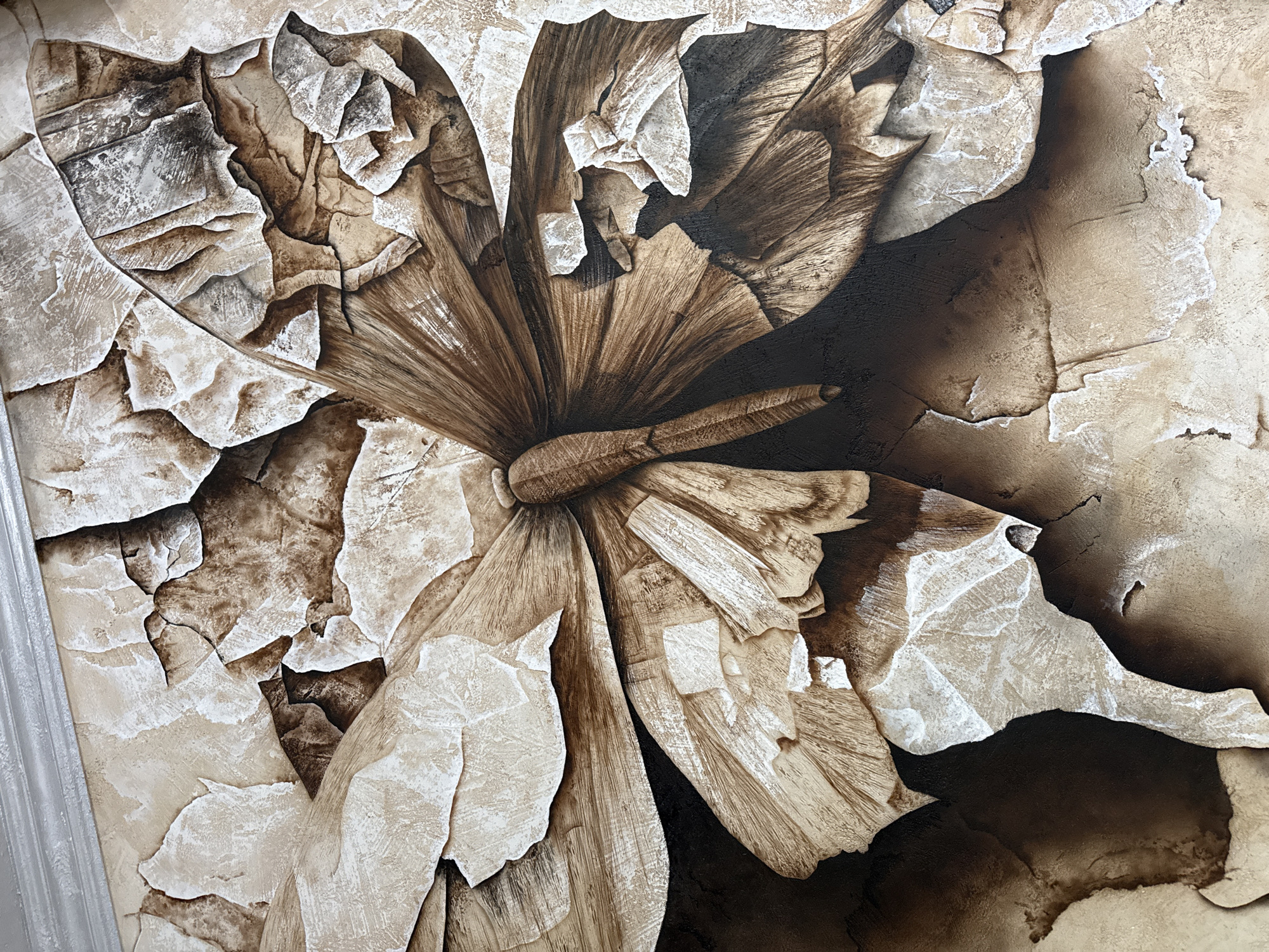 The image shows a close-up of a textured wall covering featuring a stylized floral design in shades of brown, beige, and white. The design gives the impression of a flower or blossom constructed from peeling or cracked surfaces. The background is darker brown, adding depth and contrast. A strip of light-colored framing is visible along the left edge of the image. There is no information about the location in Burgas, Bulgaria, people, subjects, actions, interactions, or the environment.