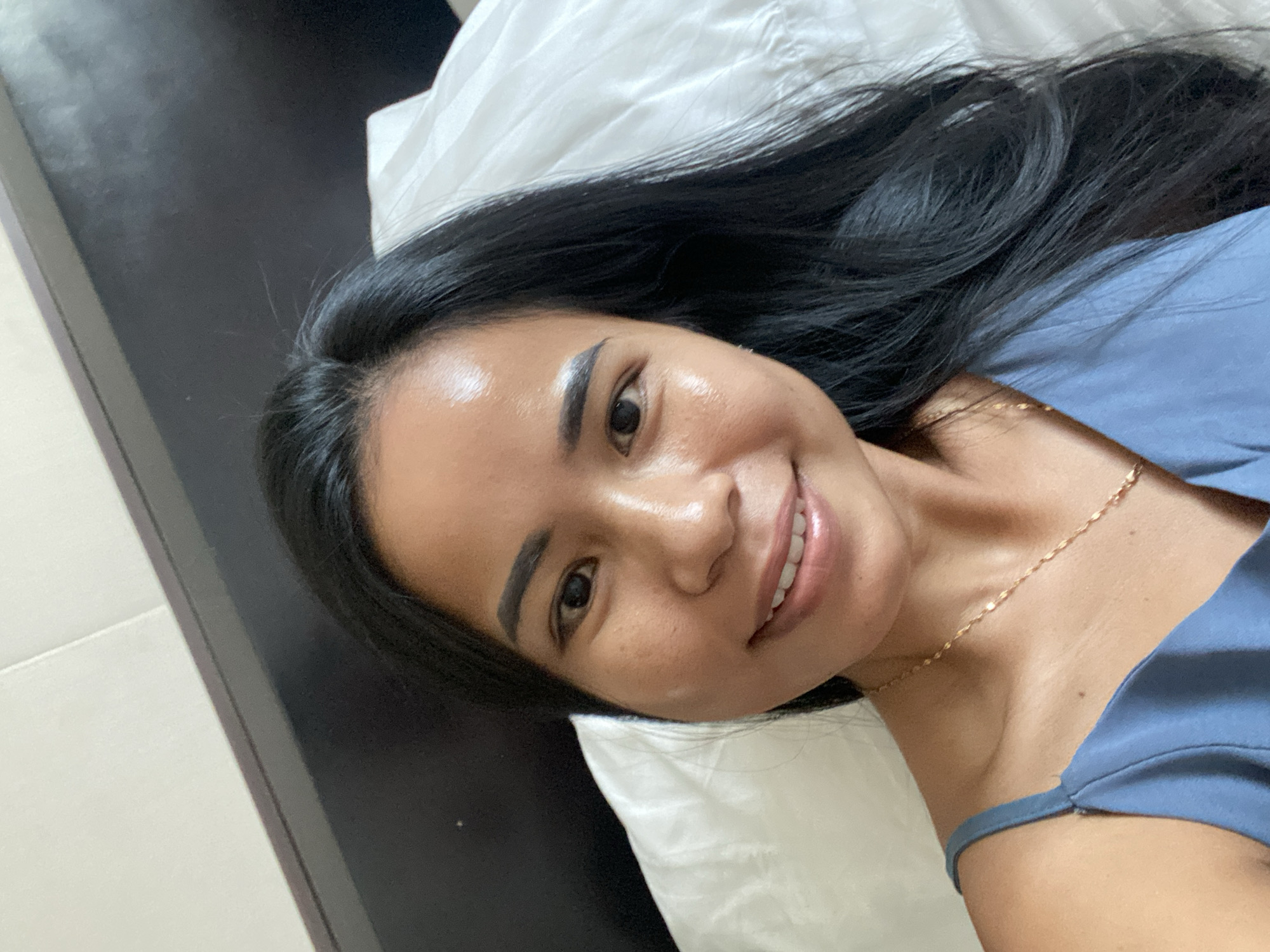 A woman with long, dark hair is lying on a white pillow. She is wearing a light blue top and a gold necklace. She is smiling at the camera. The background is dark. The image was likely taken indoors. It is not possible to verify the location without additional information.