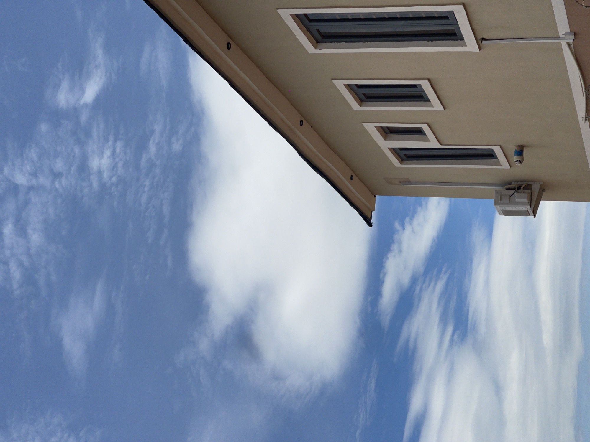 The image presents a low-angle, upward-looking view capturing the upper portion of a modern building against a partly cloudy blue sky. This scene is located in Rafin Bauna, Nigeria.
The sky, occupying the upper two-thirds of the frame, displays a varied array of clouds. Patches of clear, vibrant blue are interspersed with numerous white clouds, ranging from wispy, streaky formations suggestive of cirrus to broader, more diffuse cumulus-like structures. Some darker, greyish cloud formations are also present, indicating areas of greater density or shadow.
The building, rendered in a light, warm beige or tan stucco-like finish, occupies the lower right section. Its dark-tiled roofline features a prominent light-colored (cream or off-white) fascia and soffit, along which three circular recessed lights are visible. Below the roofline, a series of windows with matching light-colored frames punctuate the beige wall. From left to right, a tall, narrow window is followed by a smaller, roughly square-shaped window, and then another tall, narrow window. The window panes themselves appear dark, possibly tinted or shuttered.
On the far left side of the building, a white outdoor air conditioning condensing unit is mounted on the wall below the leftmost window. Associated white piping and electrical conduit are visibly connected to the unit and extend vertically downwards along the wall. Further right, below the small square window, a cylindrical object with a blue cap protrudes horizontally from the wall, appearing to be a vent or utility connection. Additional white conduits run vertically and horizontally along the building's exterior, indicating utility installations. A white trim band marks the bottom visible edge of the building's facade. No people or animals are present in the image.