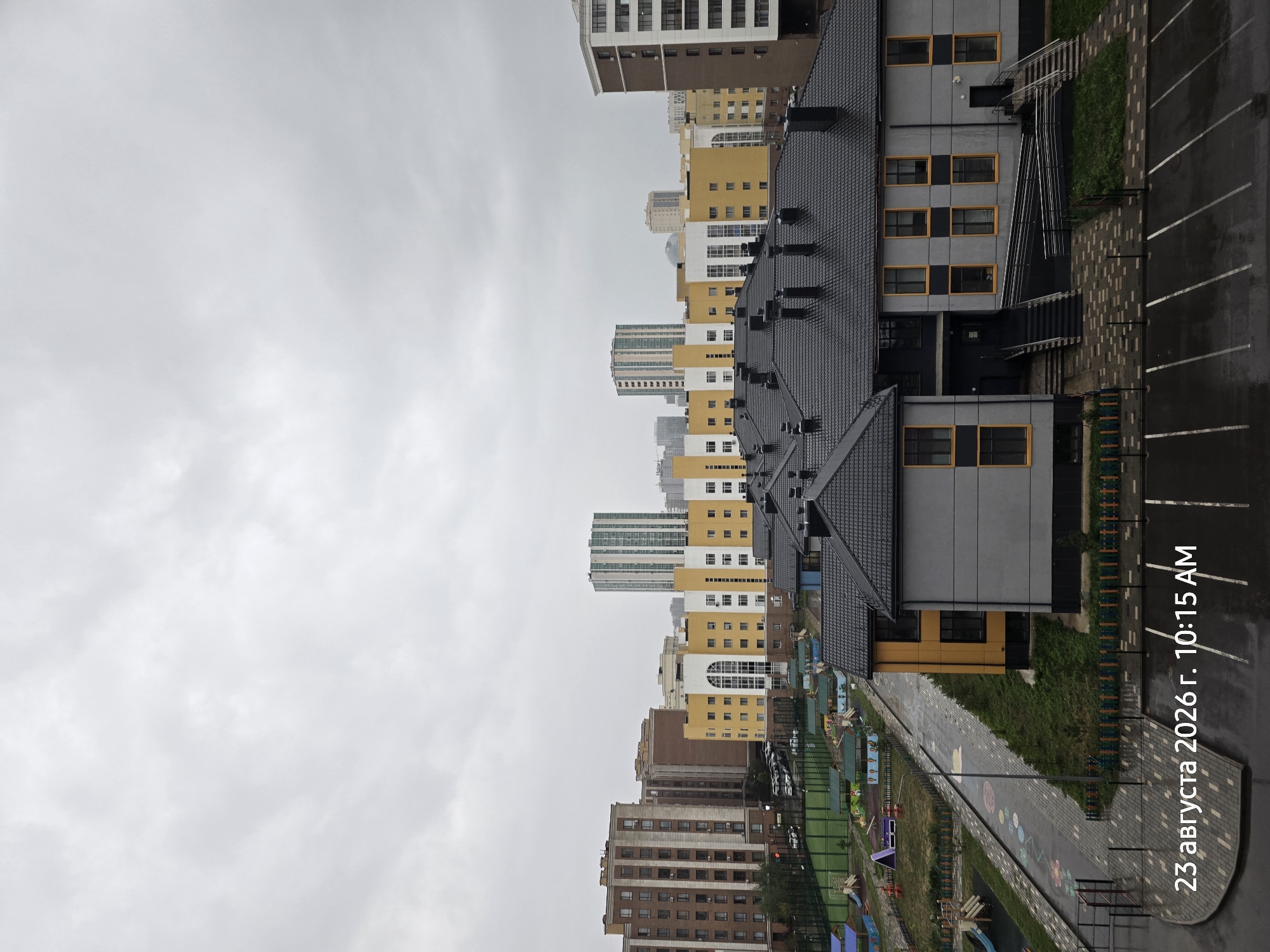 The image depicts a cityscape with multiple residential buildings and a playground under a cloudy sky. In the foreground, there is a low-rise building with a dark gray roof and gray walls, featuring yellow-framed windows. A paved area with parking lines is visible in front of this building, which appears wet, suggesting recent rain. Adjacent to it, a playground area with colorful equipment and green grass is seen, bordered by a fence. Further in the background, several taller residential buildings of varying architectural styles and colors, including brick and yellow facades, are present. Some of these buildings have distinctive green or white patterned exteriors. The overall environment suggests a residential district, possibly a new development, given the presence of a playground and modern building designs. The timestamp "23 августа 2026 г. 10:15 AM" is superimposed on the lower left corner of the image. While the location is stated as Astana, Kazakhstan, no specific landmarks are identifiable within the image to confirm this without external knowledge. No people are visible in the image.