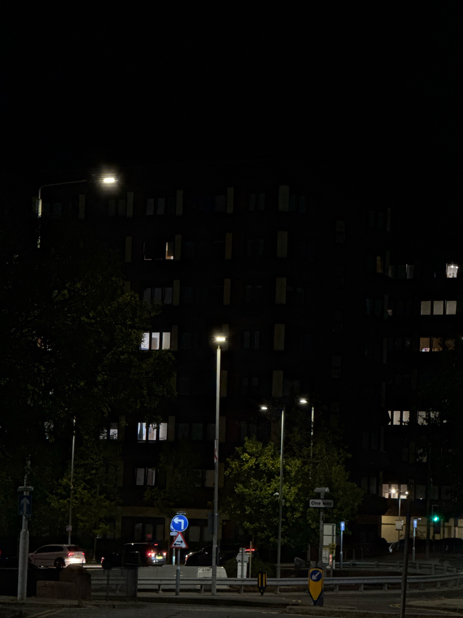 The image depicts a street scene at night in the town of Basildon, United Kingdom. In the foreground, there is a road with a white car and several traffic lights, including a green one visible on the right. There are also streetlights illuminating the area, and various road signs. Behind the road, a dark, multi-story building with illuminated windows dominates the background. Trees are present on the left and right sides of the image, some of which are lit by the streetlights. The overall impression is that of a typical urban night scene.