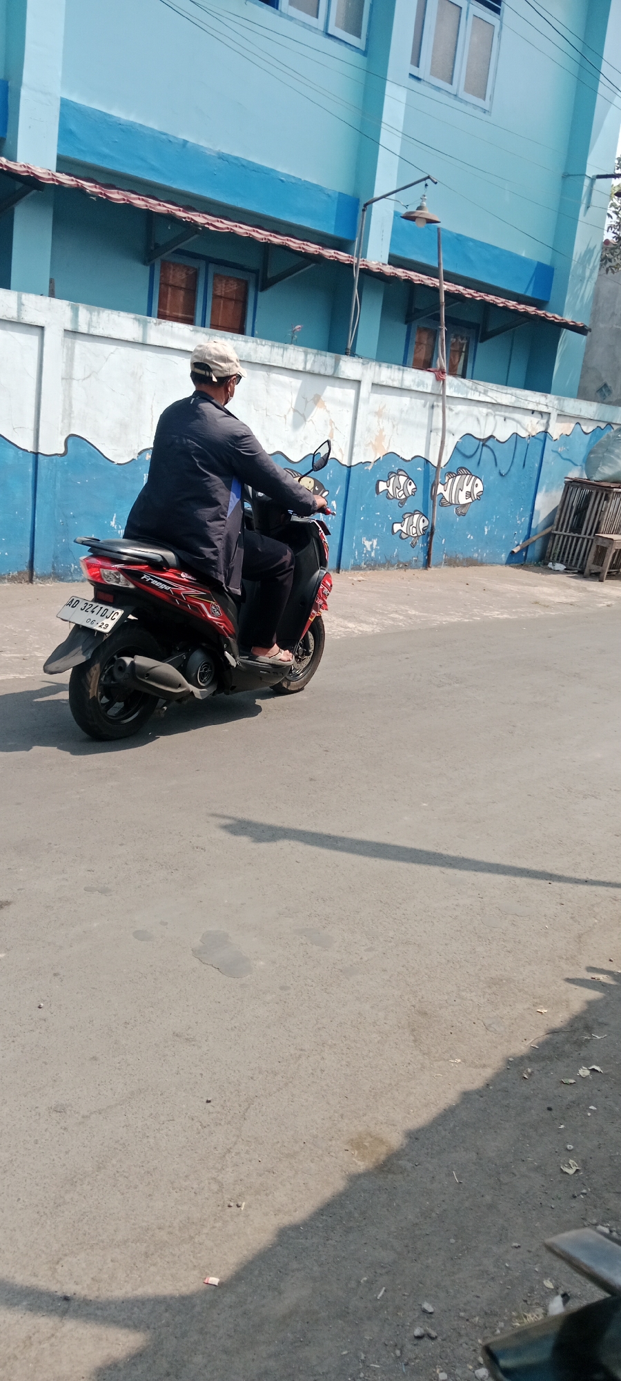 A person wearing a dark jacket, dark pants, and a light-colored cap is riding a red and black scooter. The scooter has a license plate that reads "AD 32410JC". They are on a paved road in front of a blue building with a mural of fish on the wall. The building has windows with wooden frames. A lamppost is visible on the right side of the building. The ground is asphalt, with some debris scattered on it, including cigarette butts. The lighting suggests it is daytime. This scene is located on Jalan Magelang in Purwomartani, Indonesia.