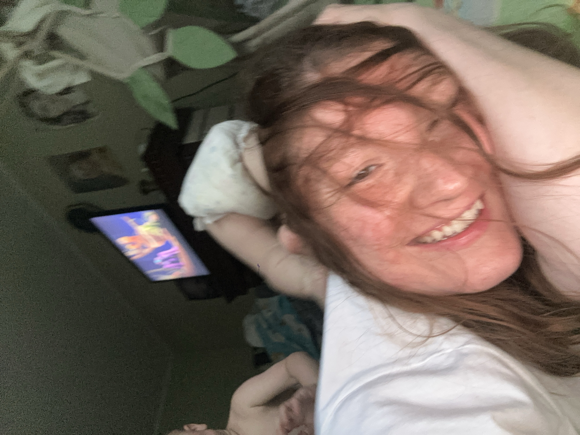 A woman with reddish-brown hair is lying on a white surface (possibly a bed) and smiling. Her hair is partially obscuring her face. A television or tablet is visible in the background, displaying what appears to be an animated program. A plant is also visible in the background. The location is not discernible from the image.