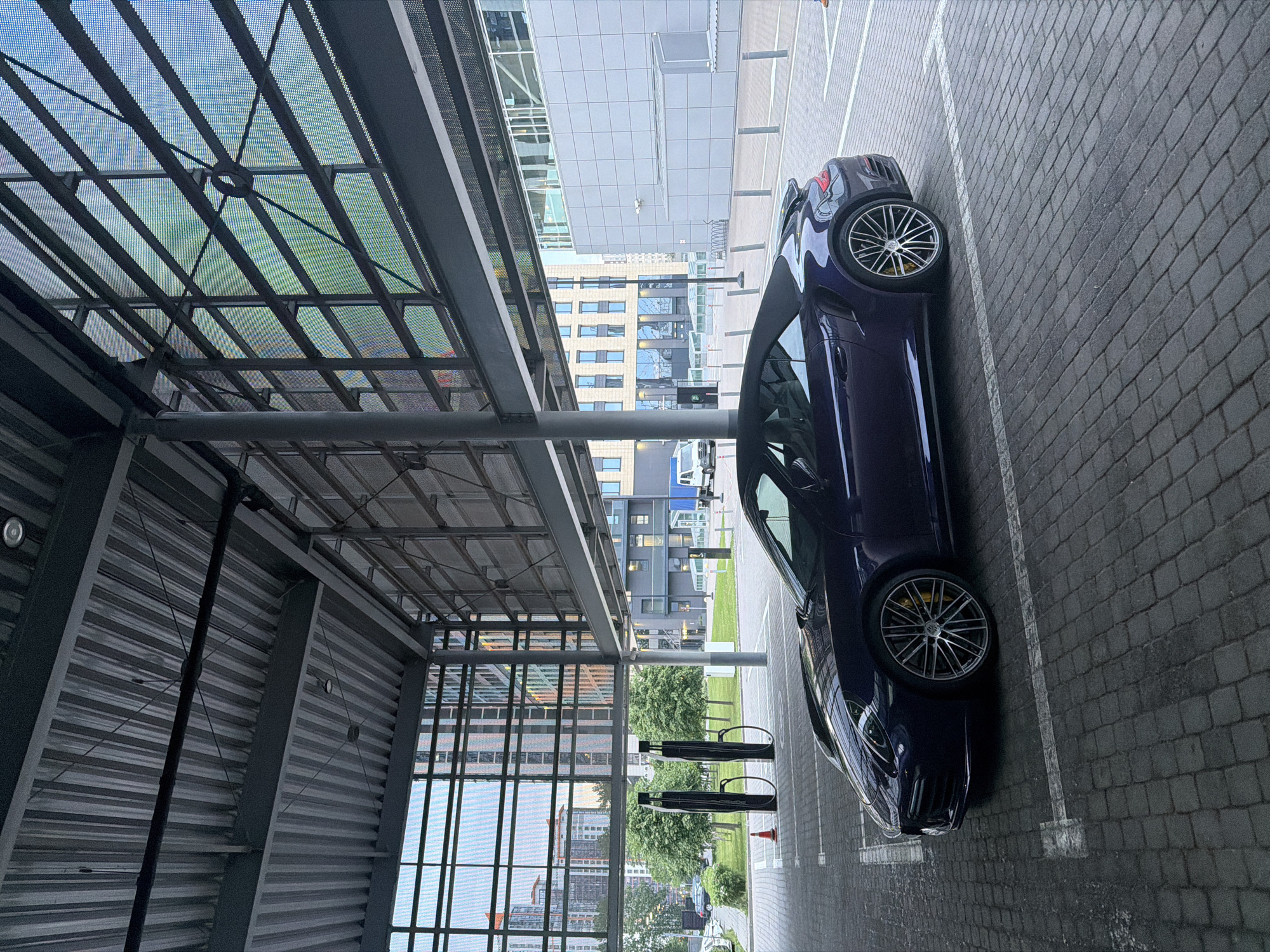 An exterior, eye-level, full shot captures a dark purple convertible sports car parked in a paved lot under a modern, covered structure. The car, a Porsche, is positioned in a marked parking space, angled slightly towards the left of the frame. Its convertible top is down, revealing the interior. The car's wheels are prominent, showcasing silver rims and yellow brake calipers.
The surrounding environment is urban. The covered structure above the car has a corrugated metal ceiling and exposed metal beams. Large glass windows with a grid-like pattern are visible to the right, offering a view of the sky and distant buildings. To the left, more glass walls reveal a park-like area with trees, other buildings, and what appear to be two electric vehicle charging stations. The ground is paved with dark gray interlocking bricks. In the background, several multi-story buildings, likely residential or office complexes, are visible, some with reflective windows. A white van is parked in the distance. The lighting suggests it is daytime, possibly overcast. There are no people visible in the scene.