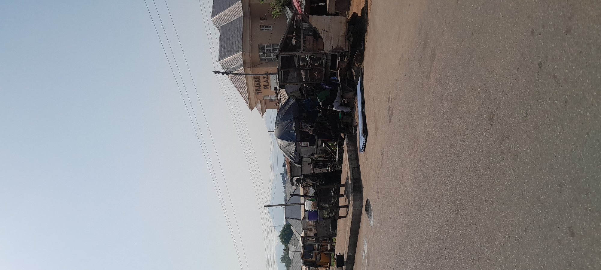 The image depicts a street scene in Jalingo, Nigeria, with a focus on a small marketplace. In the foreground, a paved road is visible. Behind the road, a raised concrete platform serves as the foundation for several market stalls.
To the left, a yellow motorized tricycle, commonly known as an "okada" or "keke," is parked. Further along the platform, a dark-colored umbrella provides shade over a stall where several people are gathered. The stalls themselves appear to be constructed from wood and corrugated metal, with some appearing makeshift.
A light brown, two-story building with a grey roof is situated in the background, bearing a sign that reads "YIWARE PLAZA." The building has multiple windows and an entrance. In front of the plaza and the stalls, power lines are strung across the sky.
Several individuals are present in the marketplace. Some are standing or sitting within the stalls, engaged in what appears to be commercial activity. One person is seen sitting on a bench in front of a stall. Another individual, wearing a dark garment, is standing near a table with containers.
The environment suggests a typical urban setting in Nigeria, with a mix of permanent structures and temporary market setups. The sky is clear and bright, indicating daytime. The overall impression is one of a bustling local market, integrated into the urban fabric of Jalingo.