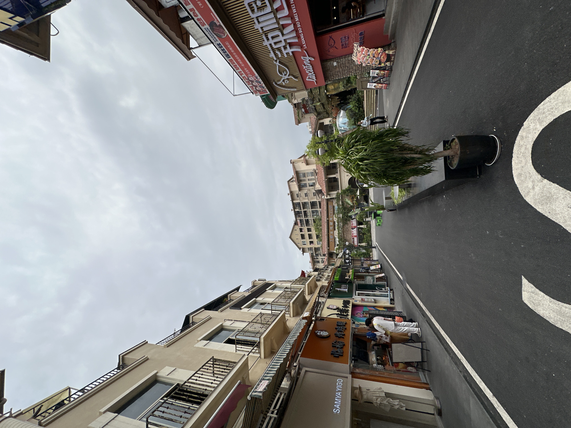 Here's a detailed description of the image:
**Location:** The image shows a street scene in the city of Chongqing High-tech Zone, China. The street is lined with commercial buildings, restaurants, and shops.
**Environment:** The sky is overcast, suggesting a cloudy day. The street is paved with asphalt and has white lane markings. There are trees and planters along the street.
**Buildings:** The buildings are multi-story, with a mix of architectural styles. Some have balconies with metal railings. Storefronts are visible at street level with various signs and awnings.
**People:** A person wearing white clothing is observed near one of the storefronts, likely working or interacting with the business.
**Objects:** Signs in both Chinese and possibly Korean (due to the restaurant name "회식 불고기 뷔페") are present, indicating the types of businesses operating in the area. There are planters with trees placed along the edge of the street.
**Specific Details (from the OCR):**
* **Restaurant Names:** The OCR identifies "LA XIANG BO BO MEI WEI N" and "55星有一家(韩国自助炭烤烤肉店)," which translates to "55 Star (Korean BBQ Restaurant)." Also, "회식 불고기 뷔페" which indicates a Korean BBQ buffet.
* **Shop Name:** "SAMYA YIGO" is listed as the name of one of the shops.
**Actions/Interactions:**
The scene shows everyday commercial activity. The person in white is likely engaged in work or customer service related to one of the businesses.