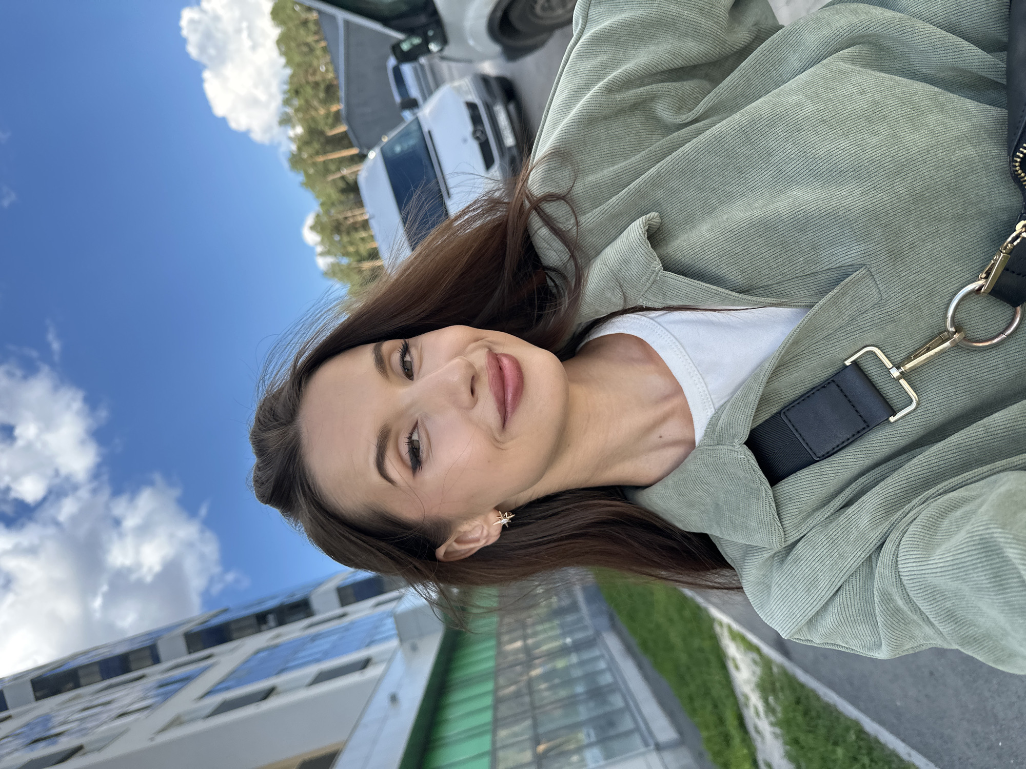 In Yekaterinburg, Russia, a young woman with long, dark brown hair and light makeup is taking a selfie outdoors. She is wearing a white t-shirt under a light green corduroy jacket and a black strap with gold hardware is visible across her chest. She is smiling at the camera, with her hair blowing slightly to her left. The background features a modern, white building with blue-tinted windows on the left, and several white vans parked on a paved area on the right. Pine trees are visible in the distance, against a bright blue sky with scattered white clouds. The ground is paved, with a strip of green grass and a concrete curb visible on the left side of the image.