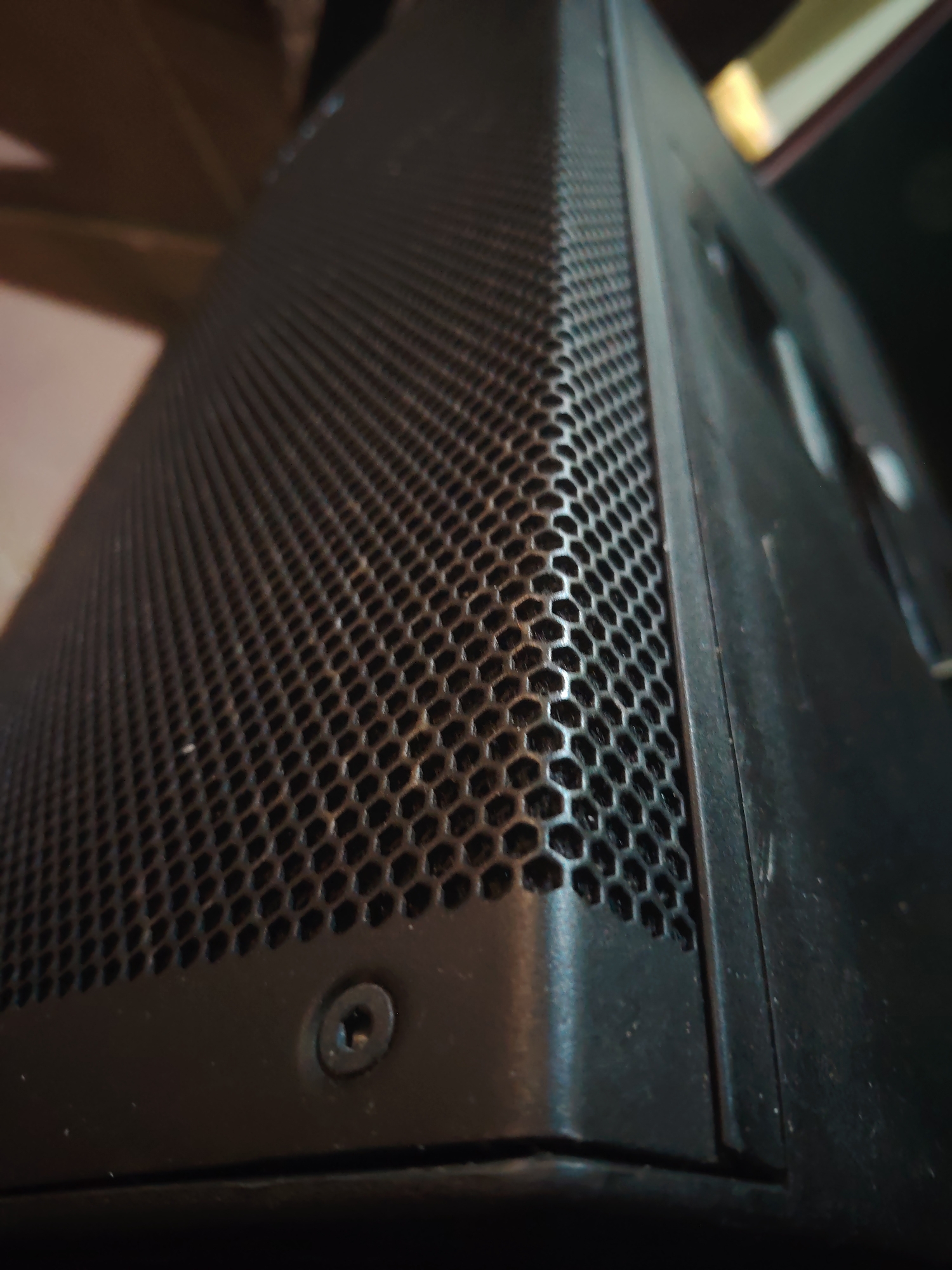 A close-up, low-angle photograph captures a section of a dark-colored electronic device, likely a speaker or a computer case. The most prominent feature is a black hexagonal mesh grille, which covers a significant portion of the device's surface. The intricate honeycomb pattern of the mesh is in sharp focus, showing individual perforations.
Below the mesh, at the bottom left corner of the frame, a single black Phillips head screw is visible, securely integrated into the device's matte black casing. The casing itself appears to be made of plastic or metal and shows some signs of use or dust. The perspective emphasizes the texture of the grille and the angular design of the object.
The background is shallowly focused and blurred, revealing indistinct shapes and muted, warm tones of brown and beige, suggesting an indoor setting. No specific activities are taking place, and there are no people or visible text. The lighting is soft, highlighting the contours and textures of the device without harsh shadows or bright reflections. The overall impression is one of a detailed examination of an industrial or consumer electronic component.