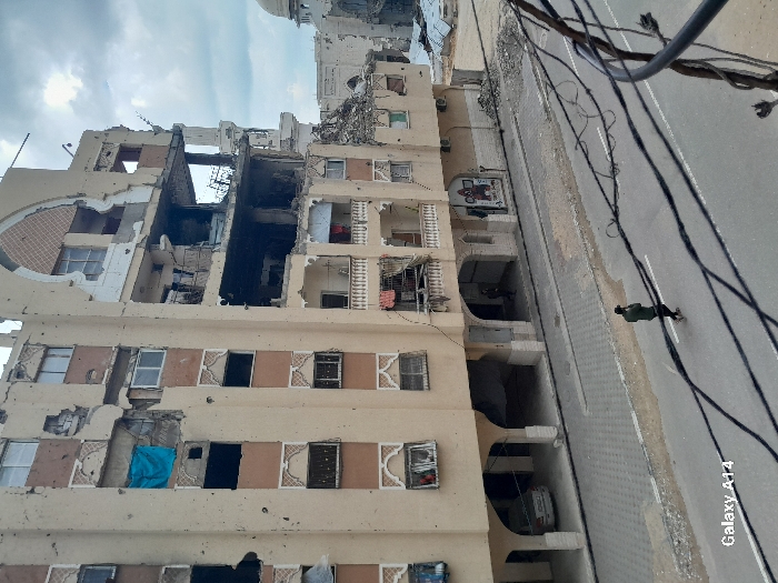The image depicts an urban scene in the city of Khan Yunis, Palestinian Territory, characterized by extensive structural damage under an overcast sky. A multi-story building, likely residential or commercial, dominates the left and center, showcasing severe destruction. Its beige facade, accented with brown panels and decorative white arch-like motifs, is pockmarked with numerous impact holes. Large sections of the upper-mid to top floors on the right side of this building are completely collapsed and blown out, revealing exposed internal structures, twisted rebar, and charred debris indicative of fire damage. A blue tarp or fabric hangs from a damaged window on an upper floor on the left, and laundry is visible draped on a lower-level balcony. The ground floor features arched open spaces; a white sedan is parked under the leftmost archway, and a dark-clad individual is partially visible under another archway further right.
In the background, to the right of the main building, further damaged structures are visible, including a mosque with a partially destroyed dome and a minaret, surrounded by scattered piles of rubble. An asphalt road with white lines traverses the lower portion of the image. A single individual, dressed in a dark green jacket and dark trousers, walks from left to right on the road, looking downwards. Multiple dark utility cables stretch across the foreground, partially obscuring the street. The scene represents an urban environment heavily impacted by conflict.