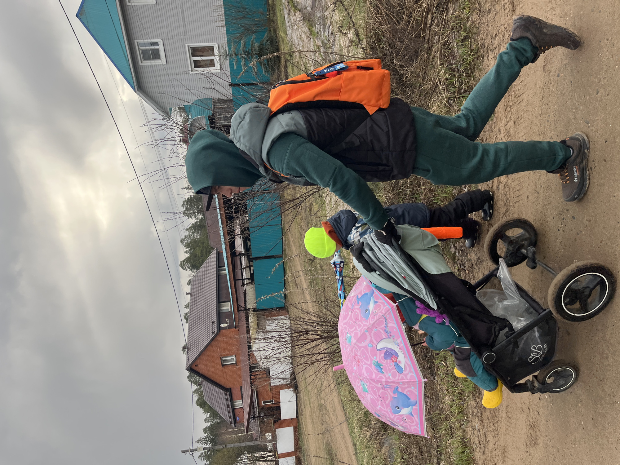 This outdoor scene, likely captured in Chaikovsky, Russia, depicts an adult and at least two children on a dirt path under an overcast sky. The image is rotated, showing subjects horizontally.
The main subject is an adult, leaning forward, dressed in a dark green hooded sweatshirt, a black and grey puffer vest, dark green sweatpants, and dark brown hiking boots (brand "Columbia" is visible on one shoe). An orange backpack is worn on their back, featuring text that appears to include "ПУТЬ" and "КПВ" in Cyrillic.
To the left of the adult, a black baby stroller, branded "Happy Baby," is present, containing a child wearing yellow rain boots. Another child, wearing a bright yellow hat, is bending down in front of the stroller. A vibrant pink umbrella, decorated with blue dolphins and other marine creatures, lies open on the ground near the stroller.
The setting appears to be a rural or semi-rural residential area. In the background, there are several houses: one is light grey with a blue roof, and another is constructed of reddish brick with a dark brown roof. Bare trees and bushes line the path and surround the houses, suggesting late winter or early spring. Overhead, power lines stretch across the cloudy, grey sky. The ground is unpaved and appears muddy. The overall atmosphere suggests a casual family outing or walk.