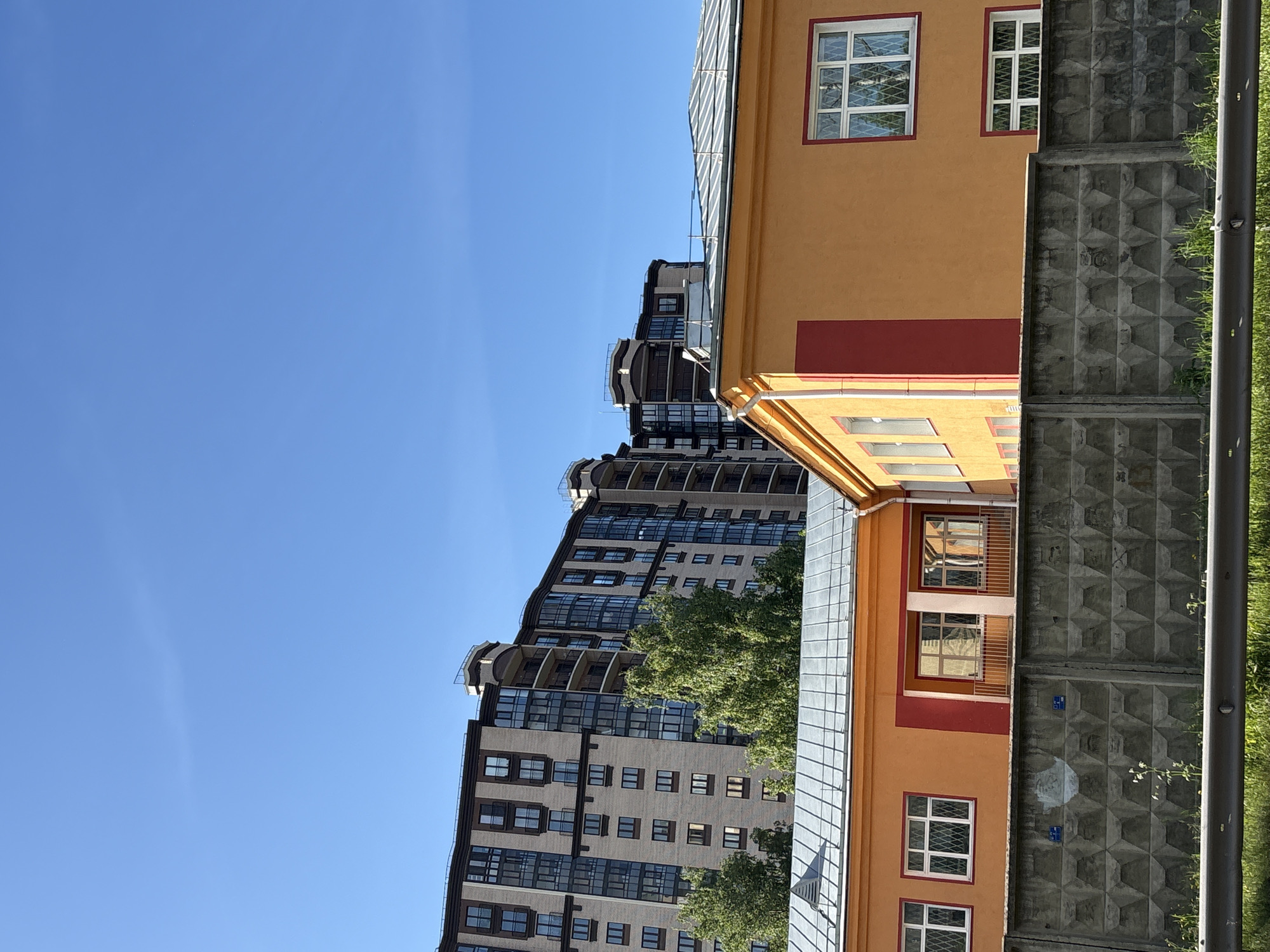 The image, rotated 90 degrees counter-clockwise, displays an urban landscape under a clear, bright blue sky. The left side of the frame is almost entirely clear sky, indicating a sunny day with no clouds.
On the right side of the image, the foreground features a two-story building with an orange-yellow facade and distinct red accents around its white-framed windows, which are adorned with white security grilles. The roof of this building appears to be made of light-colored metal.
Behind this immediate building and extending across the lower right section of the frame is a gray concrete fence. This fence is constructed from panels that have a textured, geometric pattern resembling a waffle or diamond design. Along the top of the fence and to its far right, patches of vibrant green grass and foliage are visible. A dark gray, vertical pole stands on the extreme right edge, partially hidden by the greenery.
In the background, rising above the concrete fence and the orange-yellow building, is a tall, multi-story modern residential building. It has a light brown or beige facade with numerous dark-framed windows and balconies, featuring some curved architectural elements on its upper levels. Green tree foliage partially obscures the lower and mid-sections of this taller structure.
No people or discernible activities are present in the scene. The lighting suggests it is daytime. Faint, illegible markings, possibly numbers or letters, are visible on some panels of the concrete fence, along with two small, indistinct blue square shapes that might be stickers or plaques.
