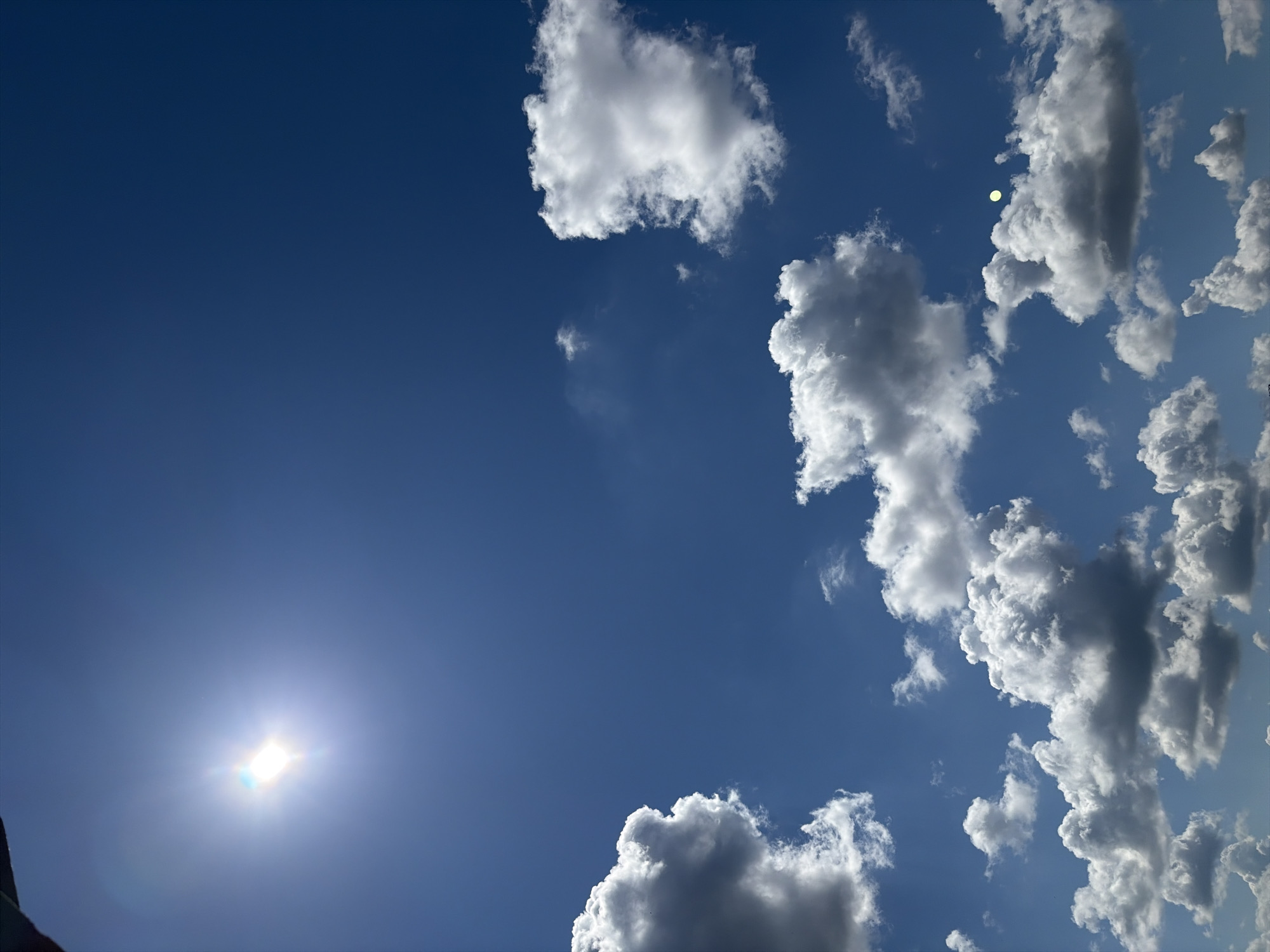 The image shows a bright blue sky with white, puffy clouds. The sun is visible in the lower left corner, appearing as a bright white light with rays emanating from it. The clouds are clustered towards the right side and top of the frame, with some scattered across the sky. The image gives the impression of a clear, sunny day. No city or location information is included in the image.