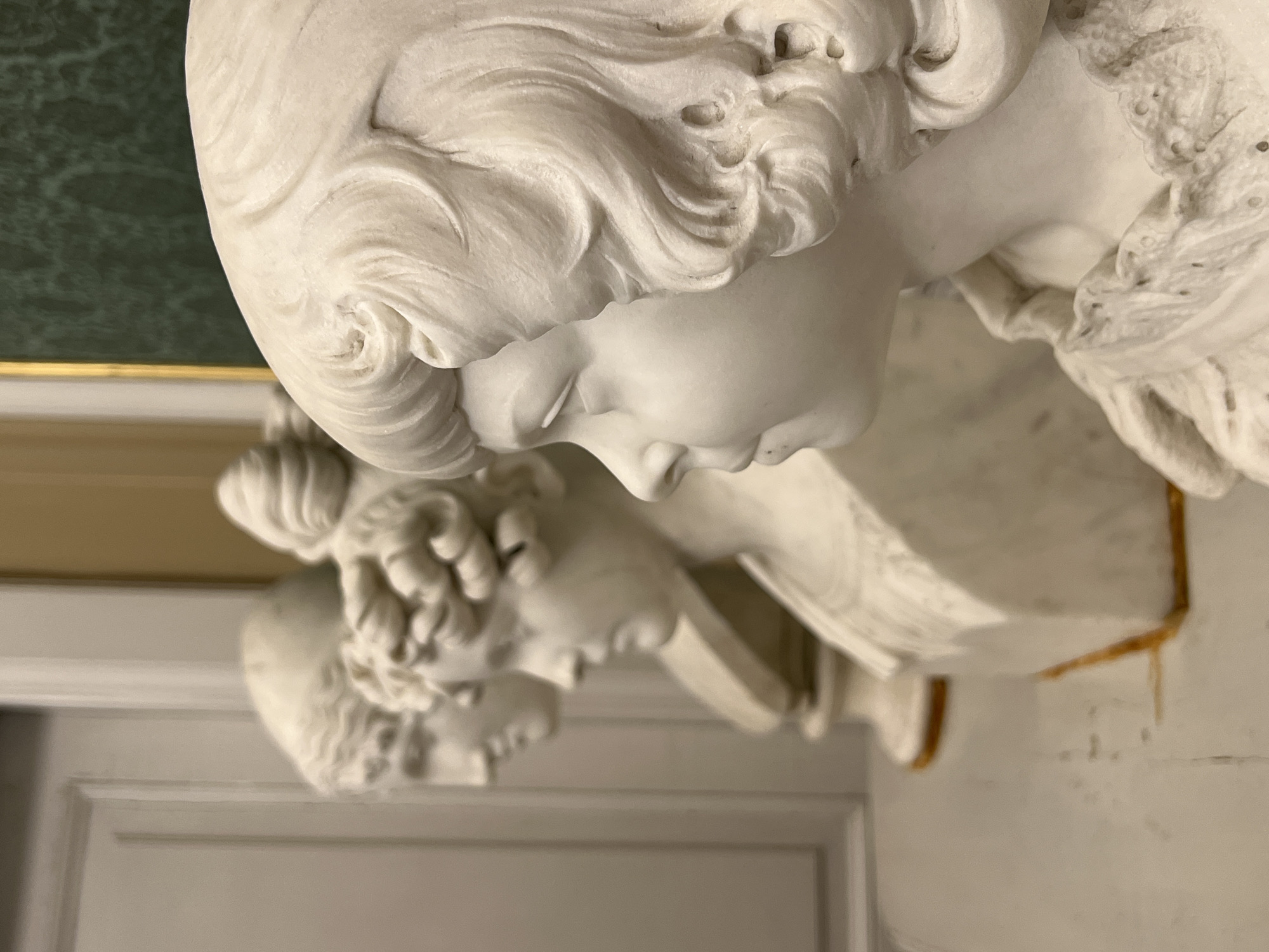 The image shows a close-up, low-angle, portrait-oriented photograph of a white marble bust of a young woman. The bust is intricately carved, featuring flowing hair that is styled in curls and waves. Her head is tilted slightly downwards, with her eyes closed and a serene, almost melancholic expression. The details of her face, including her delicate nose and lips, are rendered with a high degree of realism. The texture of the marble is visible, with some small imperfections and specks of dark material, suggesting age or perhaps the artist's technique.
Behind the bust, the background is out of focus. A sliver of a green, marbled surface is visible at the top left, with a thin gold band below it. To the right and lower, the bust is placed on a white pedestal which has some reddish-brown, possibly metallic, accents. The lower part of the image shows architectural elements that appear to be white wainscoting or paneling.
The lighting suggests an indoor setting, possibly a museum or a grand interior, with soft illumination that highlights the contours of the sculpture. The overall mood is one of quiet contemplation and classic beauty. The image is oriented vertically, with the subject appearing to be looking downwards and slightly to the left of the frame. No text is visible in the image.