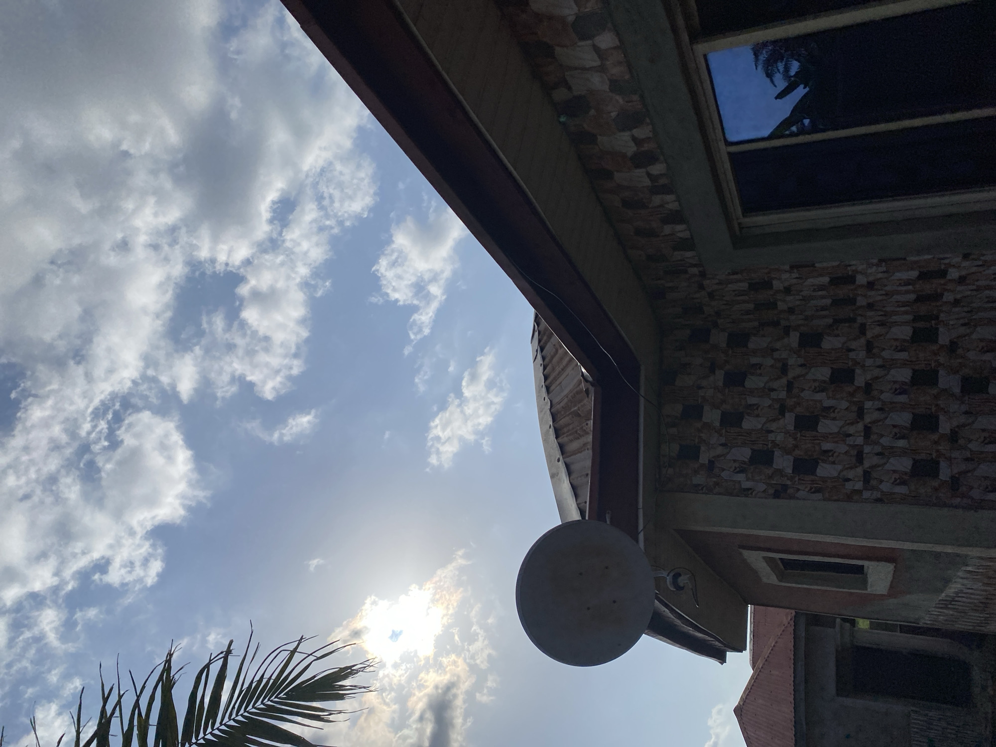 The image shows the exterior of a house in Jauro Yinu, Nigeria, with a view of the sky. The sky is partly cloudy with the sun partially obscured by clouds and a palm frond in the upper left corner. A satellite dish is mounted on the side of the house, above a window. The house has a textured exterior wall with a patterned design, and a brown roof. Another building with a red roof is visible in the background. The scene appears to be captured from a low angle, looking upwards.