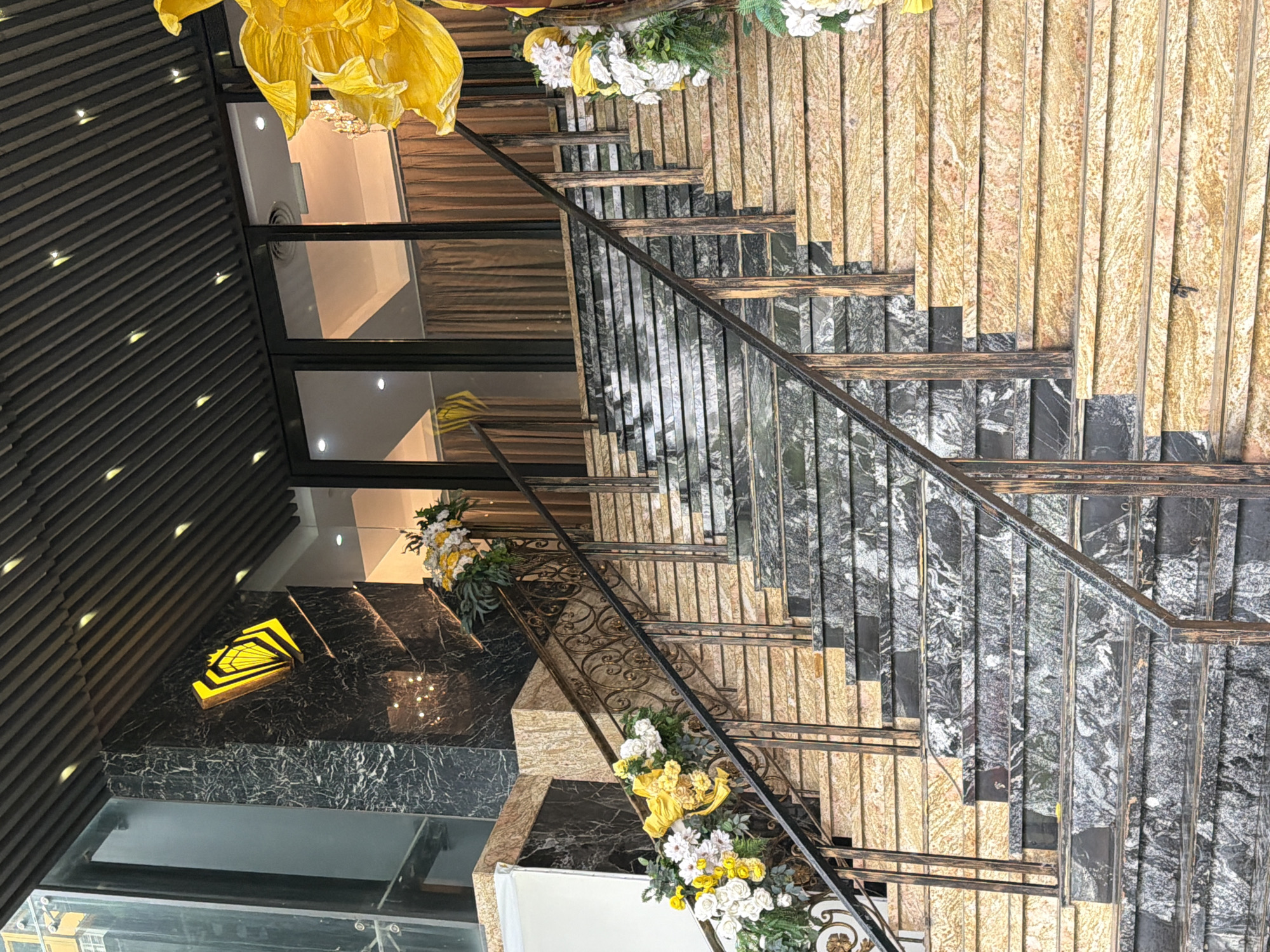The image shows a grand entrance with a wide, multi-tiered staircase leading up to a building. The staircase is made of polished marble with alternating bands of black and speckled beige stone. Ornate metal railings with intricate scrollwork line both sides of the stairs. Large, artificial yellow flowers are prominently displayed at the top of the stairs, and smaller floral arrangements in white and yellow with greenery adorn the railings on the lower steps.
Above the entrance, a dark, slatted ceiling is visible, punctuated by small, circular lights. A prominent black marble column to the left features a glowing yellow logo with a geometric design. The building entrance itself has large glass doors, with brown curtains visible behind them.
The image is taken from a low angle, looking upwards at the entrance. The overall impression is one of luxury and opulence, likely a hotel, restaurant, or event venue. The specific location is not explicitly identified in the image, but the style suggests a high-end establishment.